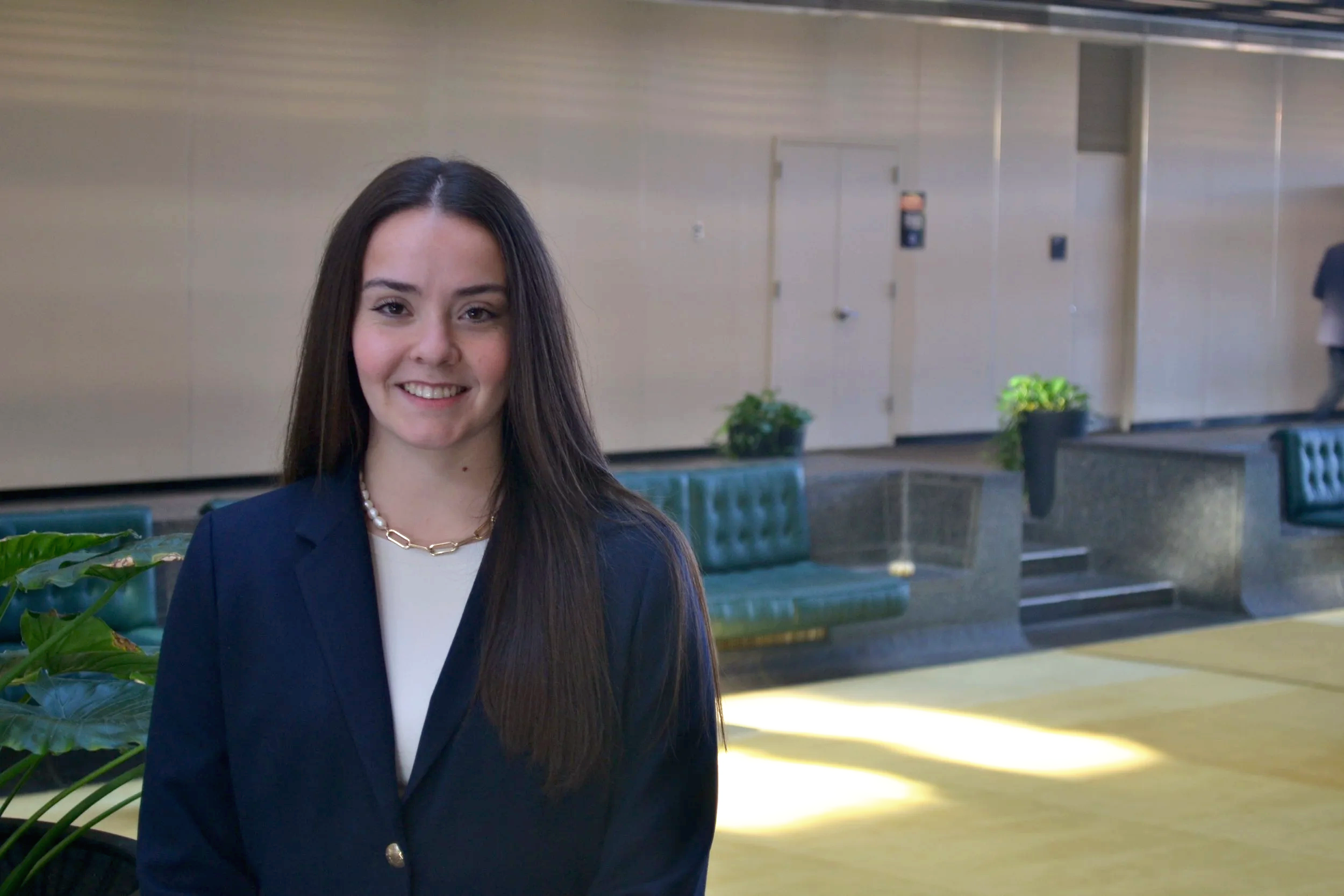 A young woman with long dark hair, wearing a navy blazer and jewelry, smiling in an indoor setting with green seating and potted plants in the background.