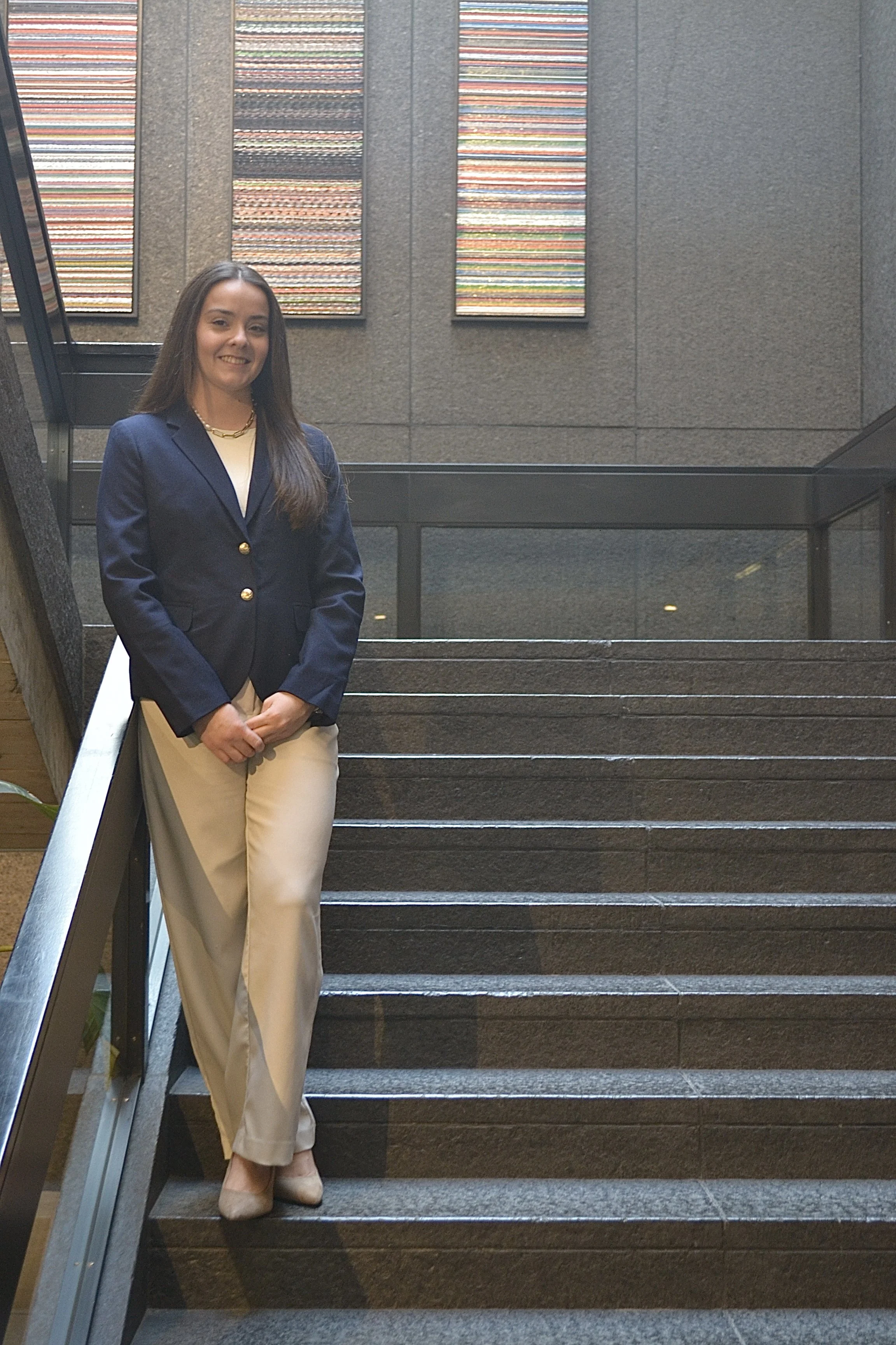 A woman standing on a staircase in a modern building, wearing a navy blazer, cream-colored pants, and beige shoes, with abstract artwork on the wall behind her.