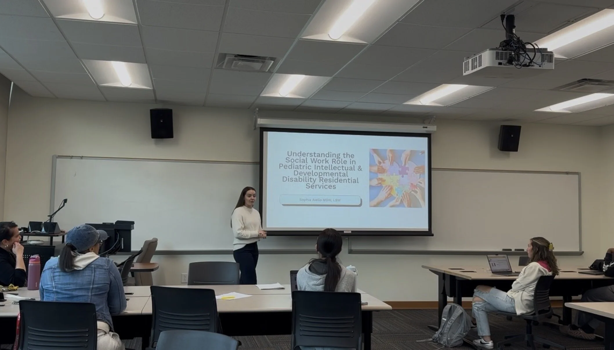 A woman giving a presentation in a classroom with a projection screen. Several students are seated at tables watching her.