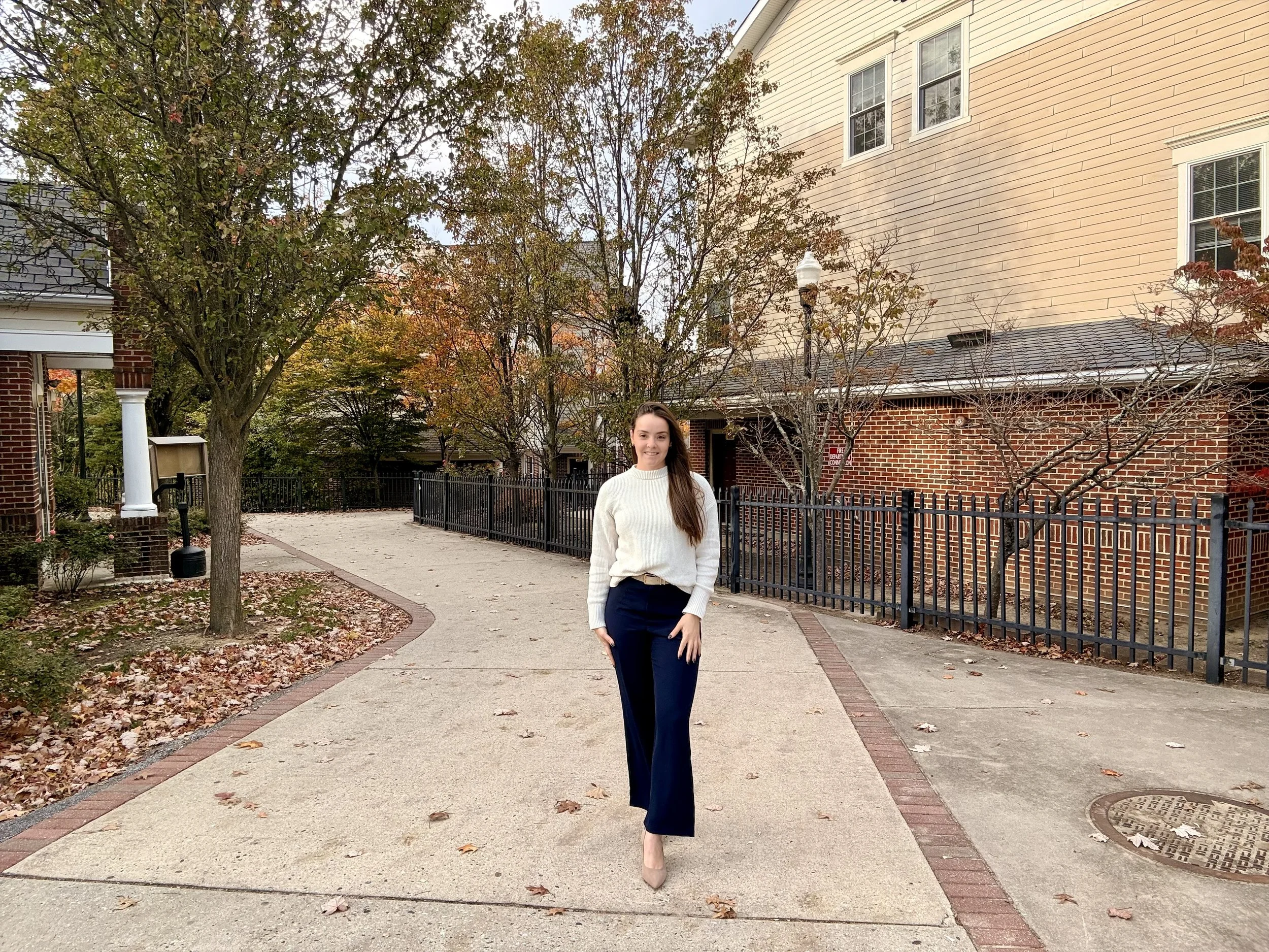 A young woman with long brown hair smiling and walking along a sidewalk in a residential neighborhood during fall, wearing a white sweater and navy blue pants.