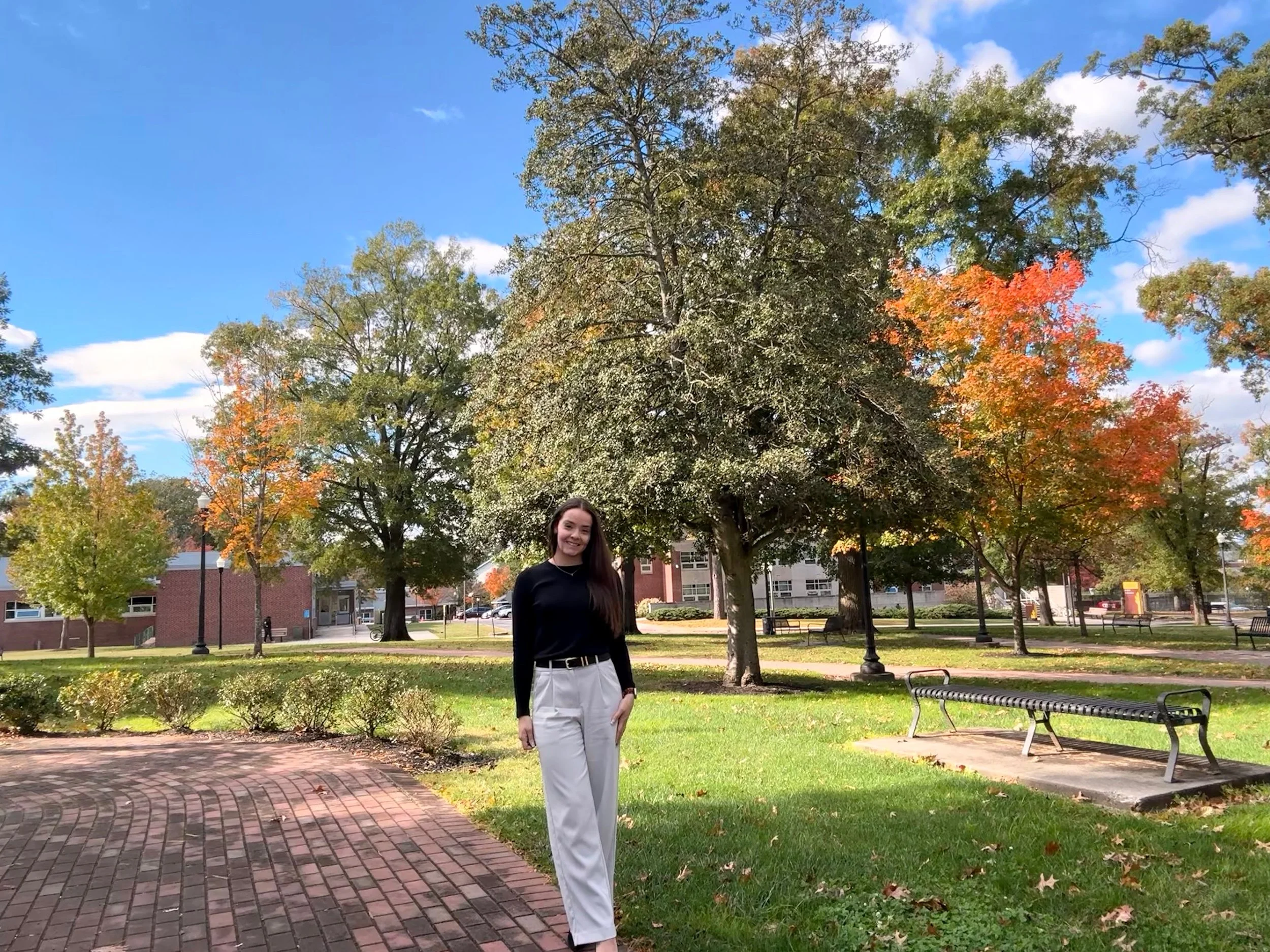A woman with long dark hair, wearing a black long-sleeve top and white high-waisted pants, smiling while standing on a brick pathway in a park during fall with colorful trees and a blue sky in the background.