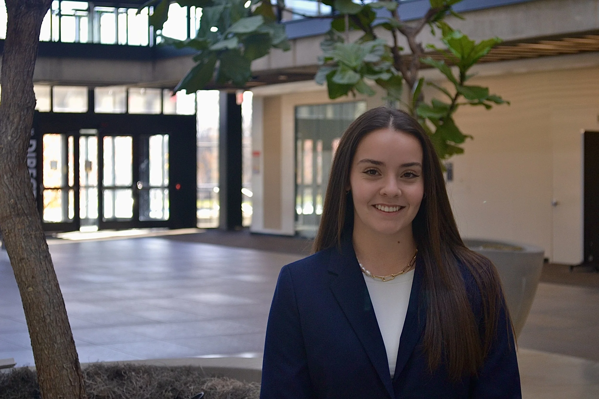 A young woman with long brown hair, smiling, wearing a navy blue blazer and a white top, standing indoors near a plant, with a modern building entrance visible in the background.