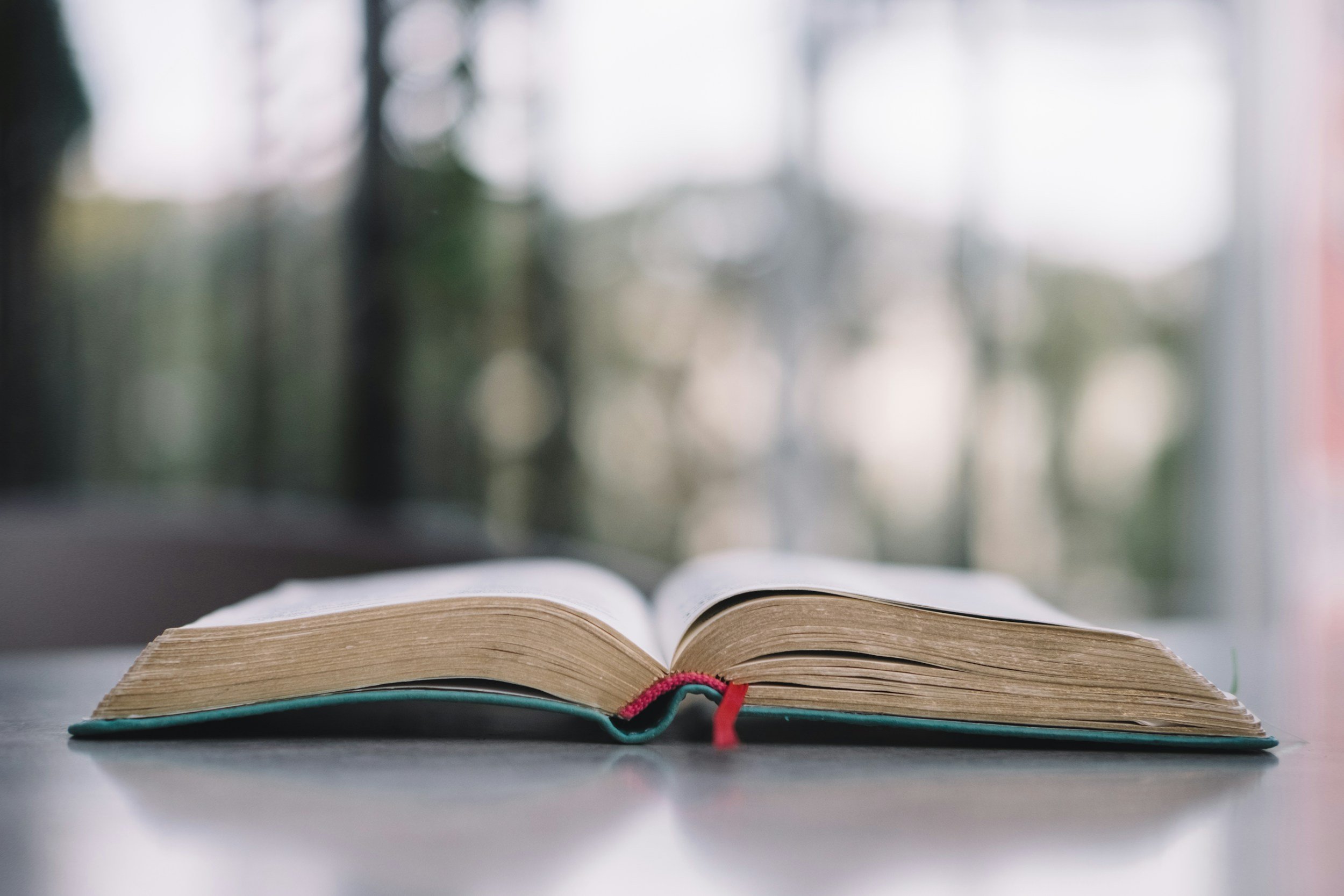 An open book with a red ribbon bookmark on a table, with a blurred window and trees outside in the background.