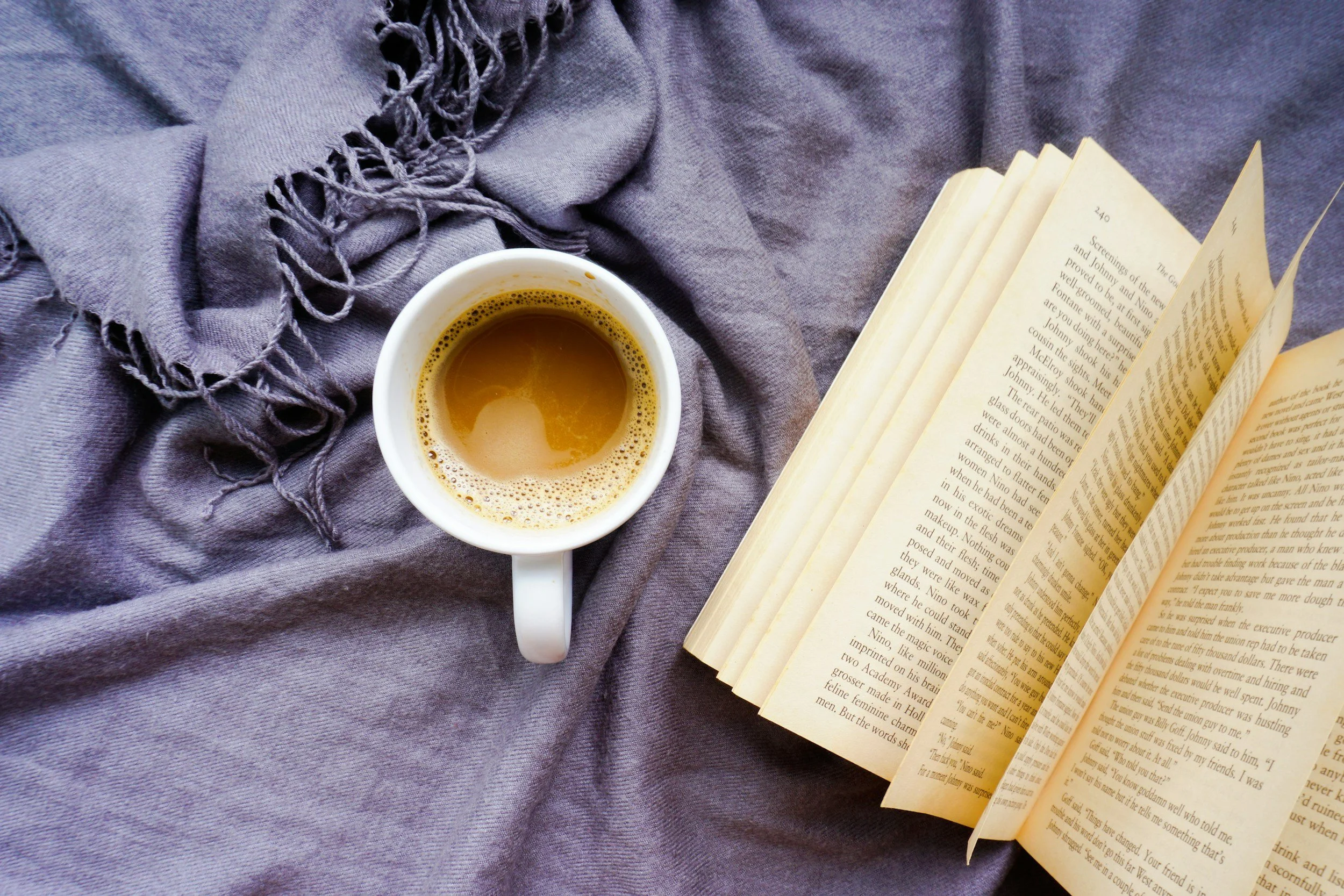 A white coffee mug filled with coffee placed on a purple fabric with an open book nearby.