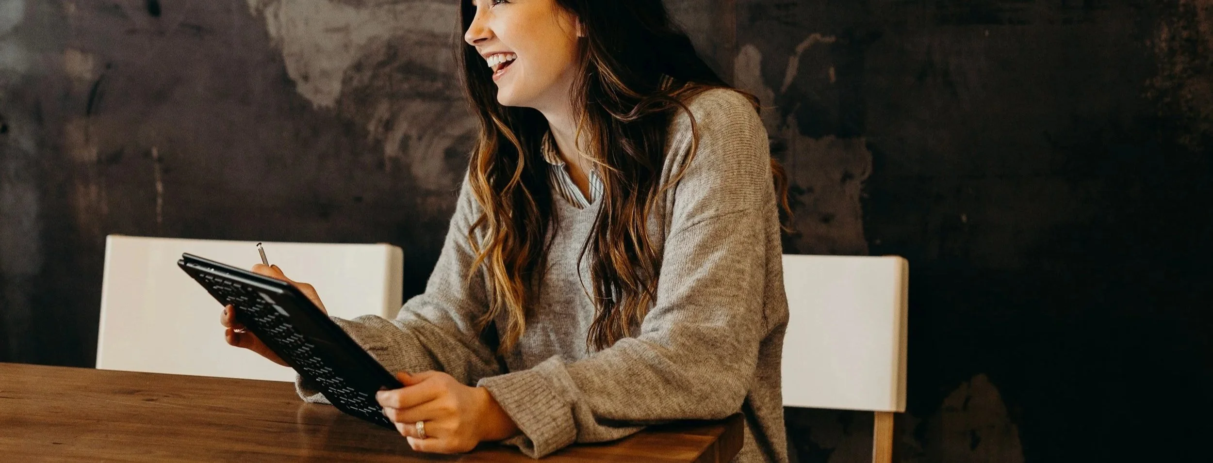 A woman with long wavy hair, wearing a beige sweater and a collared shirt underneath, sitting at a wooden table and smiling while looking at a black notebook with white text, with a dark textured wall in the background.