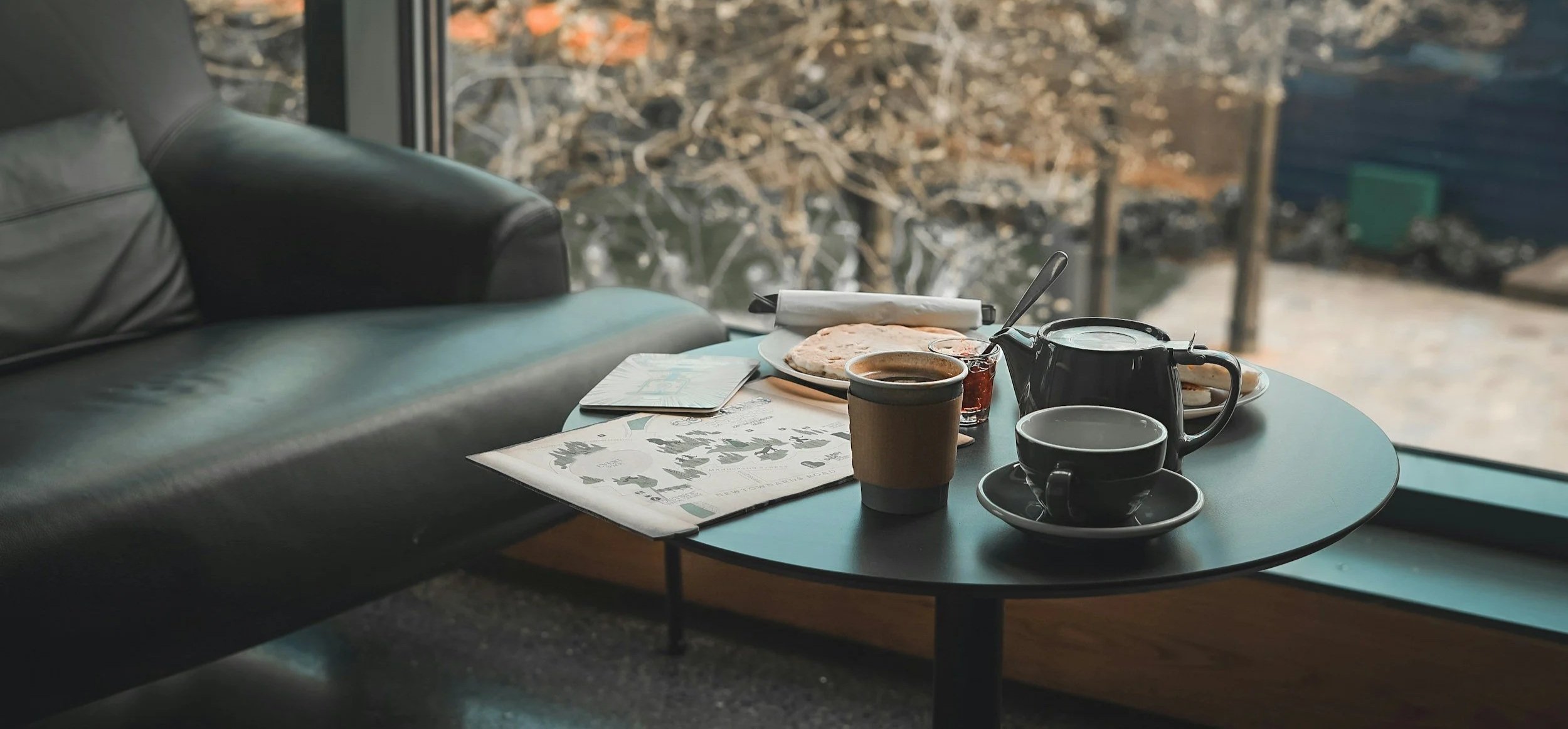 A round black table with a cup of coffee, a teapot, a cup with a saucer, a small bowl, a plate with bread or pastry, a glass of jam or syrup, and a newspaper, next to a large window with a view of trees with autumn foliage.