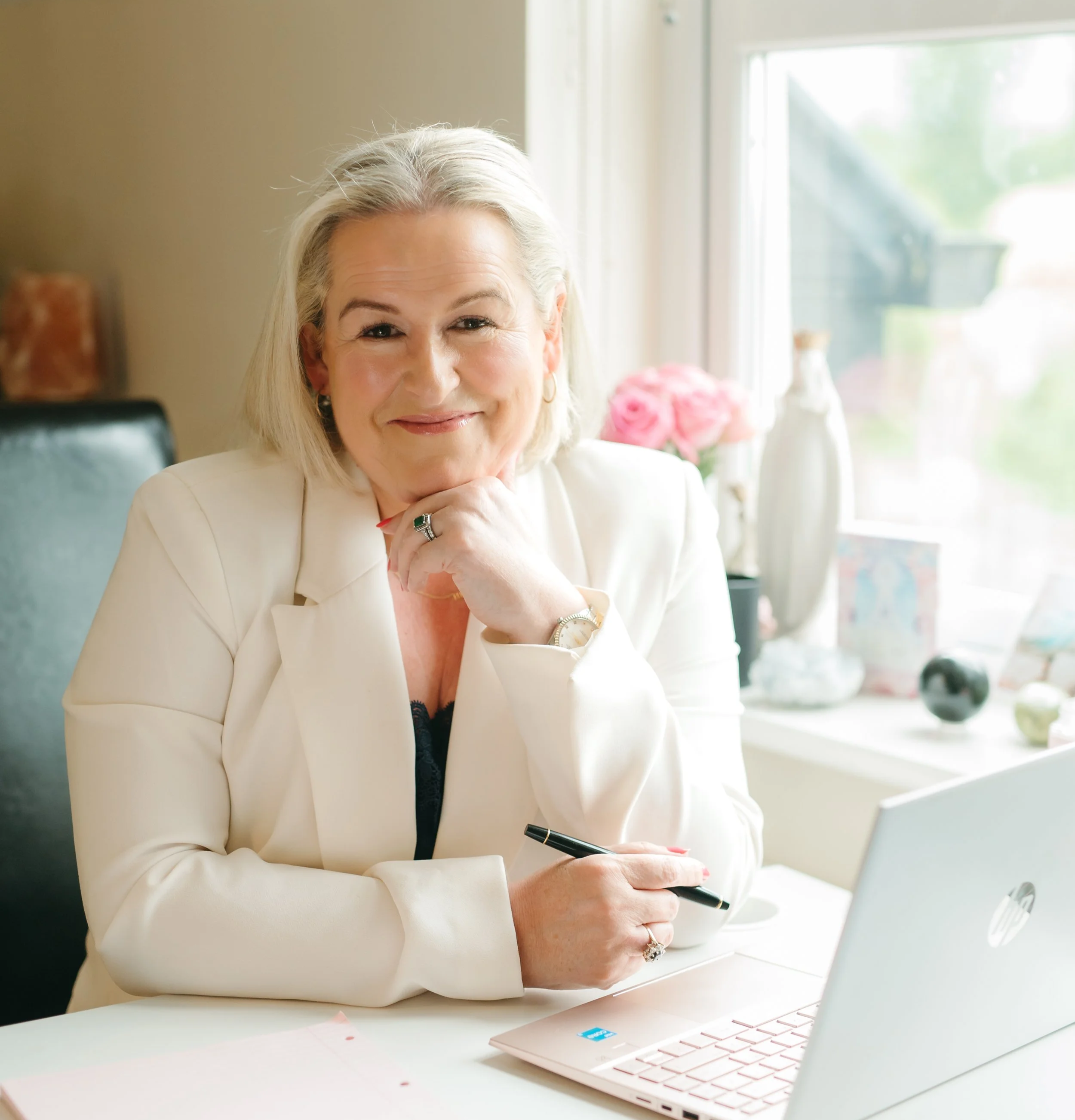 A smiling older woman with gray hair, wearing a white blazer, sitting at a desk with a pink laptop. She is holding a pen and resting her chin on her hand.
