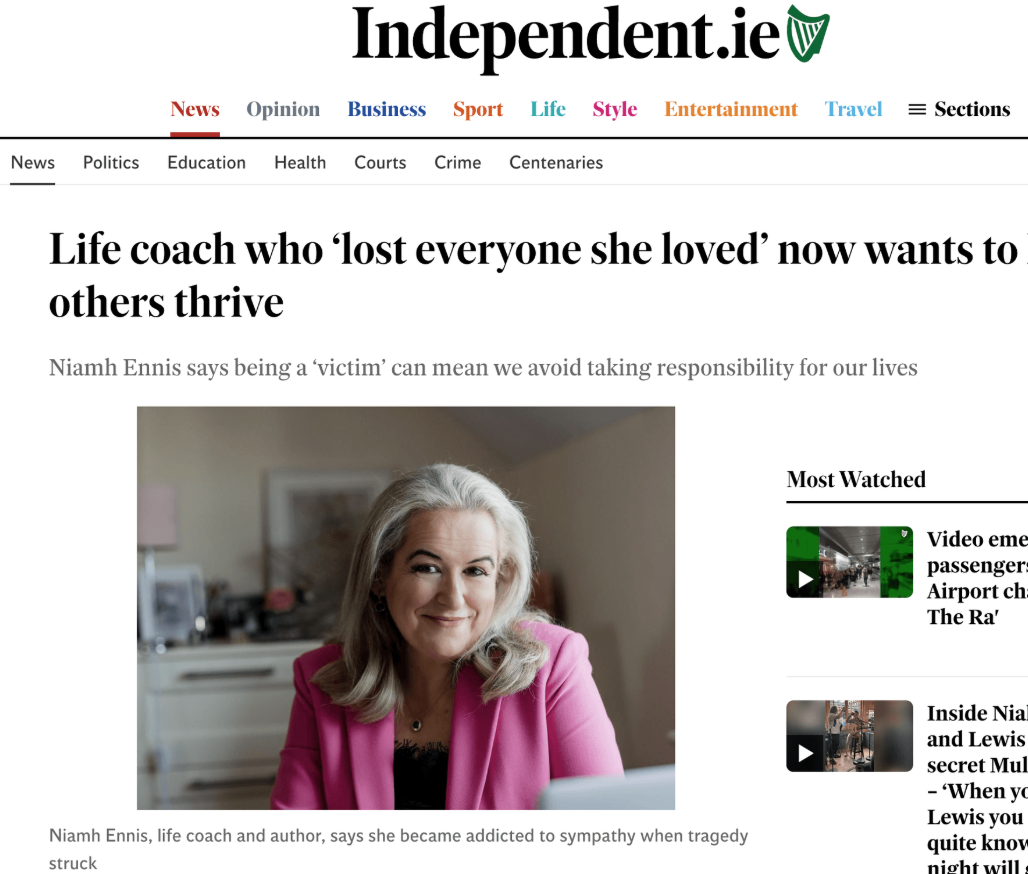 A smiling woman with gray hair wearing a pink blazer sitting indoors, with a blurred background including a framed picture and some electronic devices.