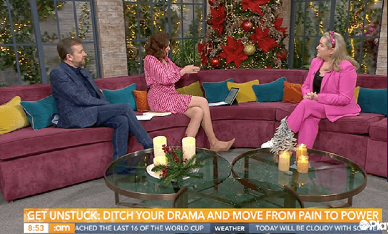 Three people sitting on a velvet red sectional sofa decorated with colorful cushions and holiday decorations, including a Christmas tree with ornaments and candles, during a daytime television talk show.