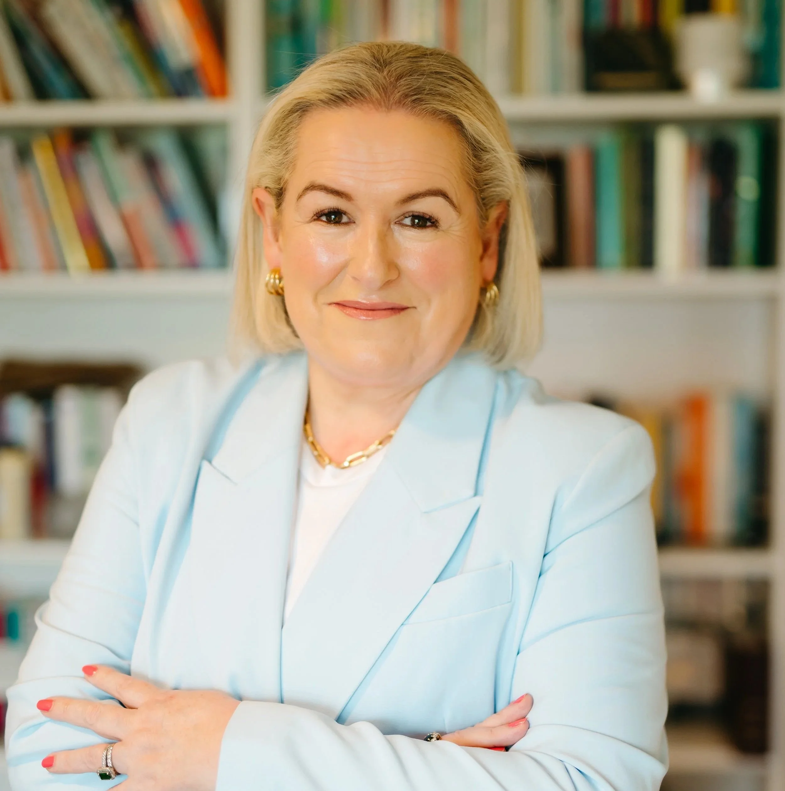 A woman with blonde hair, wearing a light blue blazer, gold jewelry, and a white blouse, smiling with arms crossed in front of a bookshelf filled with colorful books.
