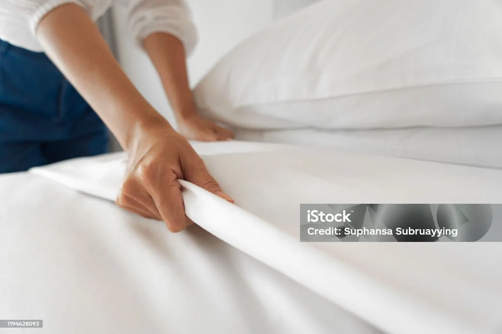 Person making a bed, tucking in white sheets.