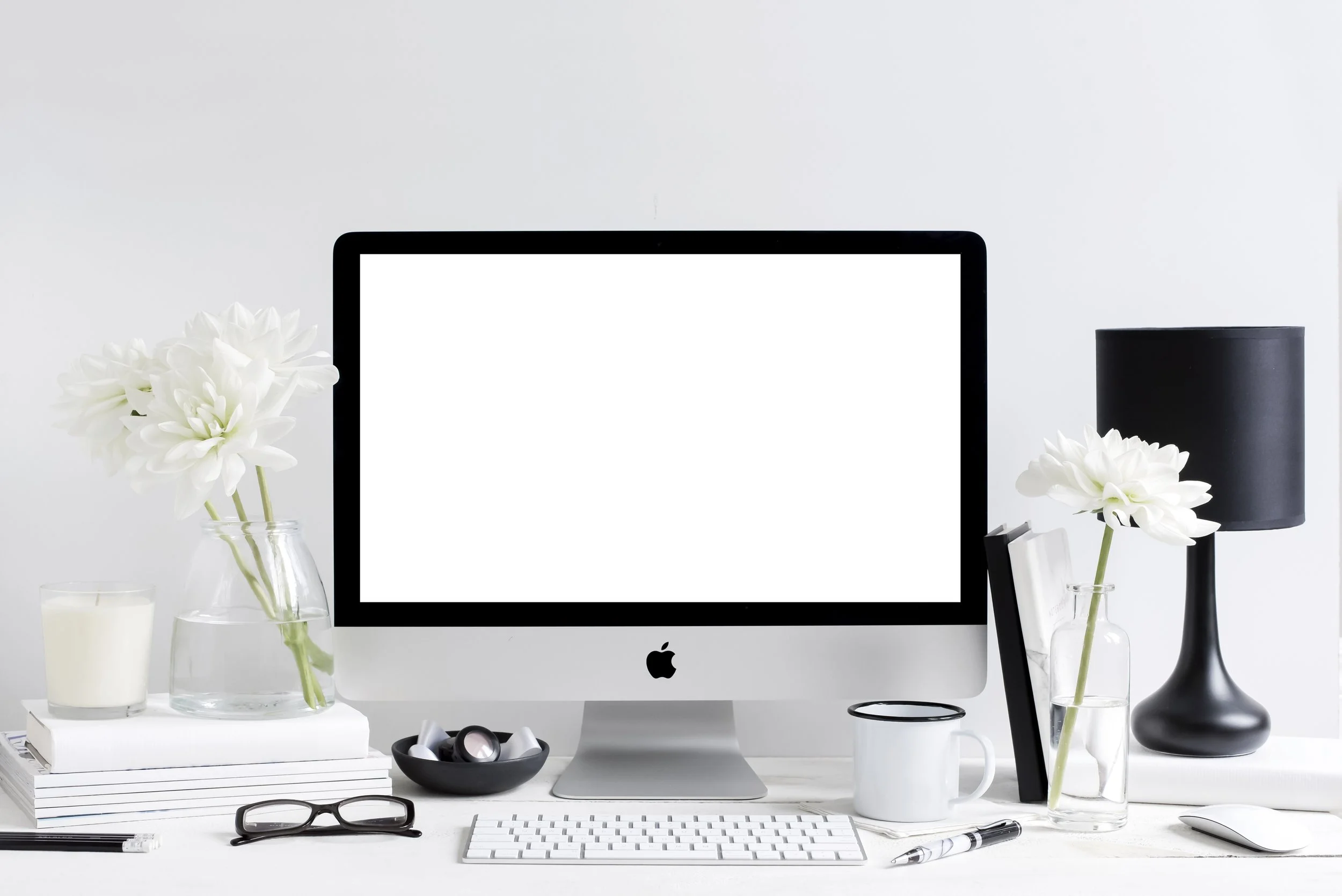 Minimalist desk setup with an Apple iMac, white keyboard, black lamp, eyeglasses, books, white daisy flowers in glass vases, candle, pen, and coffee mug.