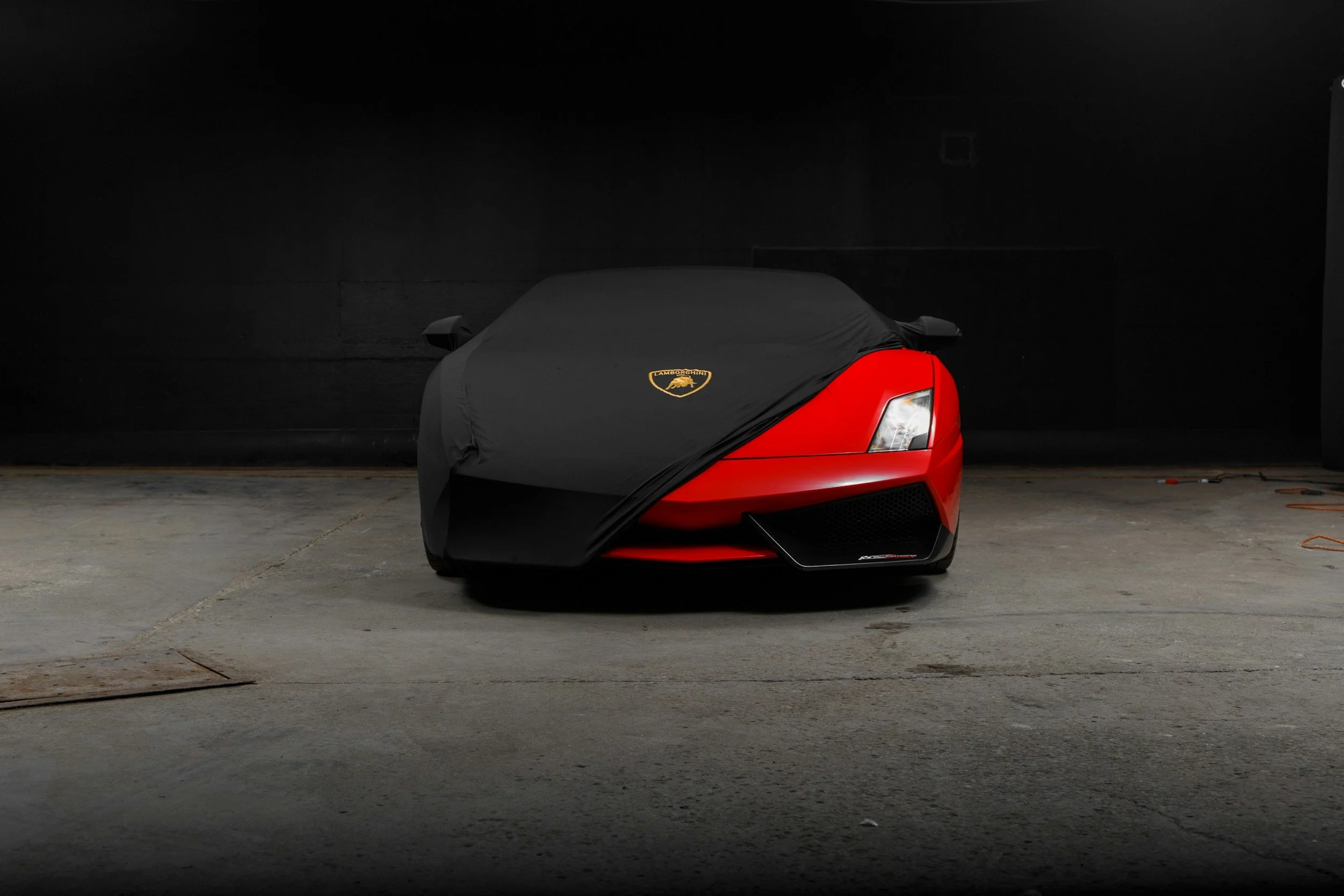 Red sports car partially covered with a black car cover in a dimly lit garage.