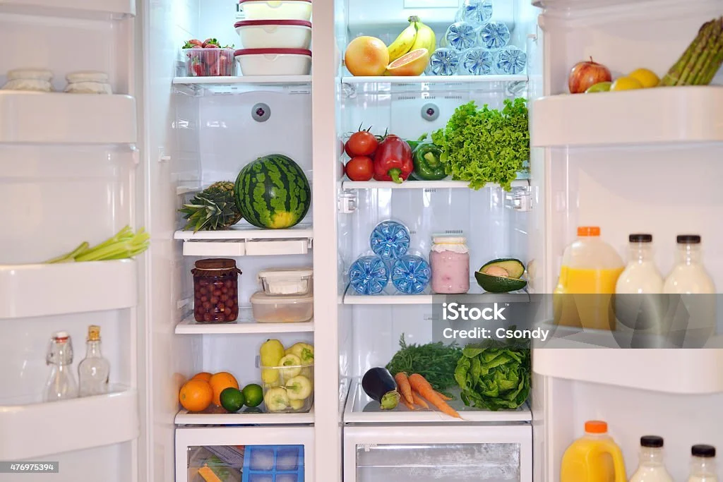 Open refrigerator filled with fruits, vegetables, and beverages. Visible items include watermelon, lettuce, bell peppers, tomatoes, bananas, oranges, carrots, celery, bottled water, milk, and orange juice.