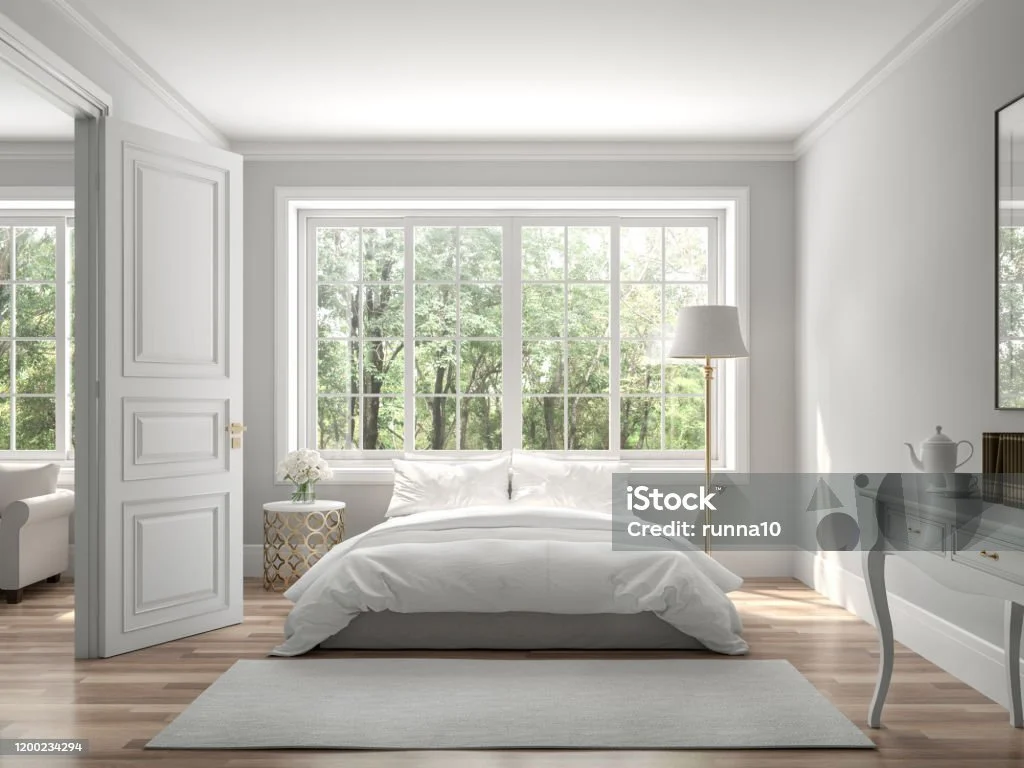 Bright, modern bedroom with white bedding, large window with a nature view, side table with flower vase, standing lamp, and gray rug. Open door to adjoining room.