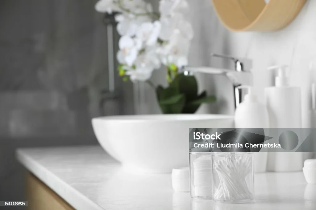 Modern bathroom counter with white ceramic sink, cotton swabs, and pump bottles. Orchid plant in background.
