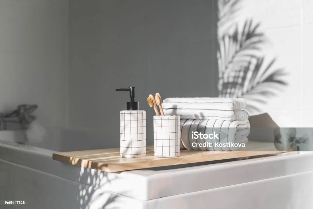 Bathroom corner with a wooden tray on a bathtub, holding a white pump soap dispenser and a container with toothbrushes. Folded towels are stacked neatly beside, with soft lighting creating leaf shadows on the wall.