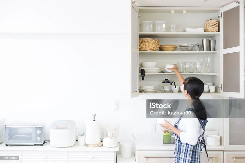 A woman organizing dishes in a white kitchen with open cabinets, containing bowls, glasses, and baskets. A countertop with kitchen appliances like a toaster and rice cooker is visible.