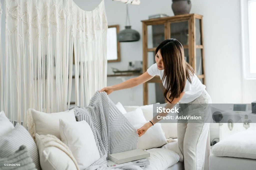 Woman arranging cushions on a couch in a cozy living room with a macrame wall hanging and light decor.