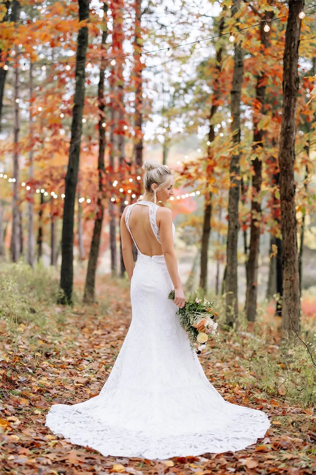 A bride in a white wedding dress holding a bouquet of flowers in a wooded area with fall foliage and string lights overhead.