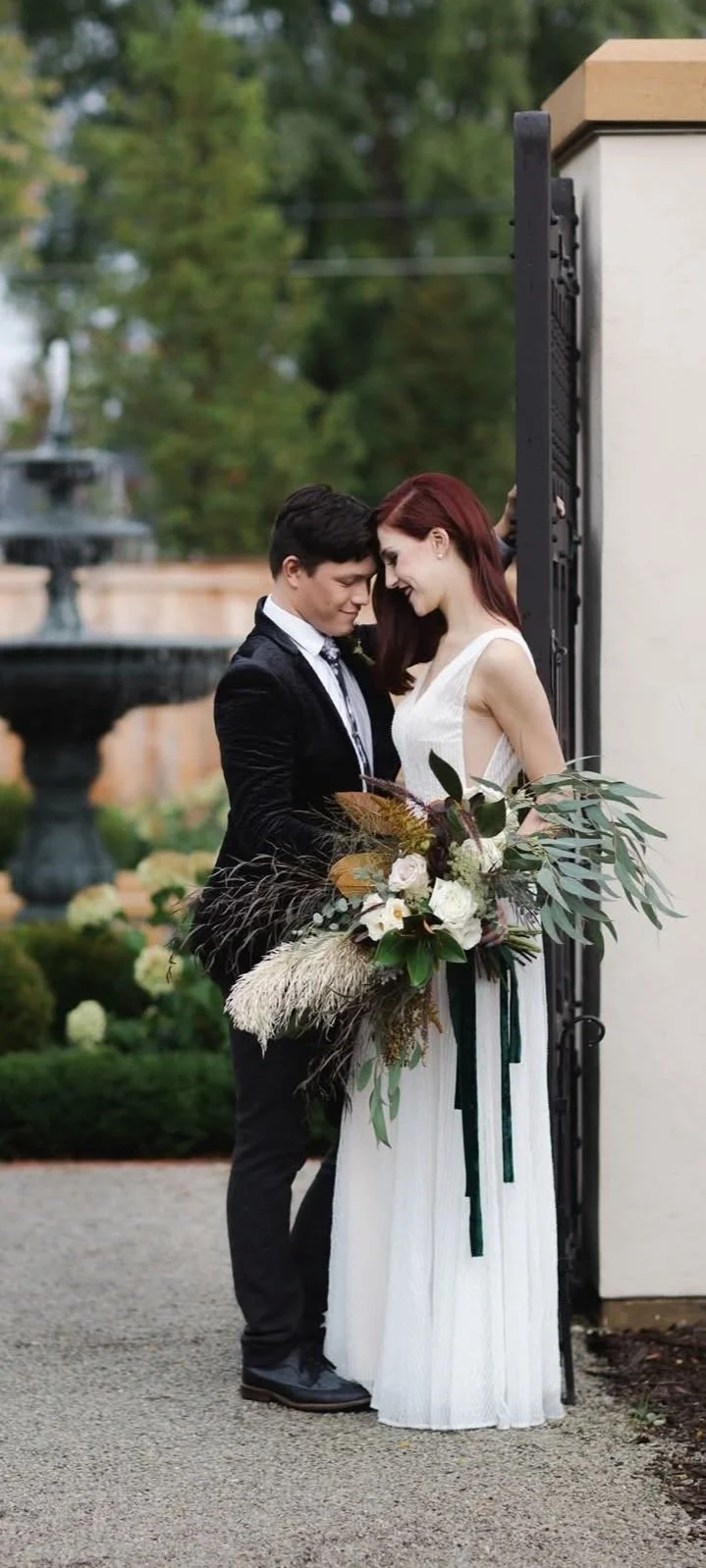 A couple dressed in wedding attire sharing a close, intimate moment outdoors, with the woman holding a large bouquet of flowers, and a fountain and lush greenery in the background.