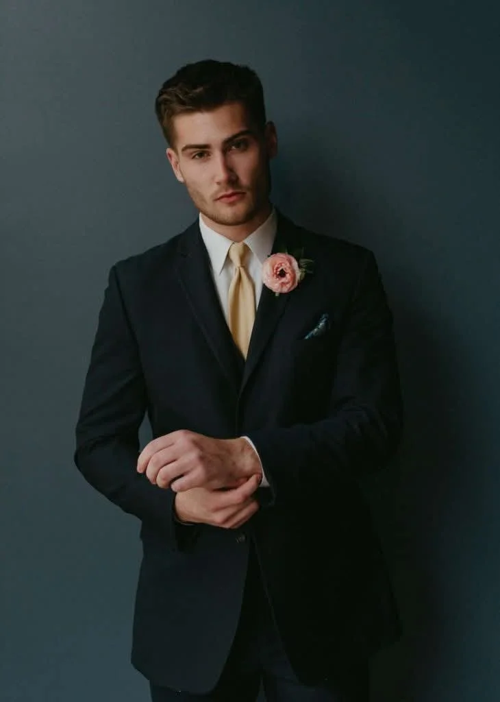 Young man in a black suit with a light-colored tie and a pink boutonniere, posing against a dark background.