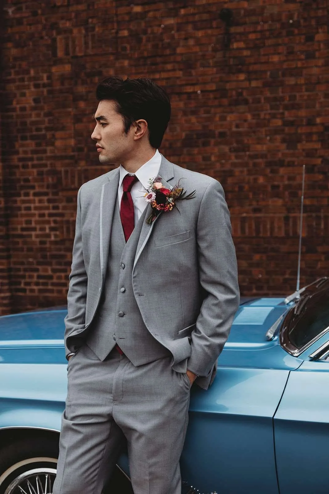 A man in a gray suit with a red tie and a boutonniere, standing with his hands in his pockets, leaning against a blue vintage car with a brick wall in the background.