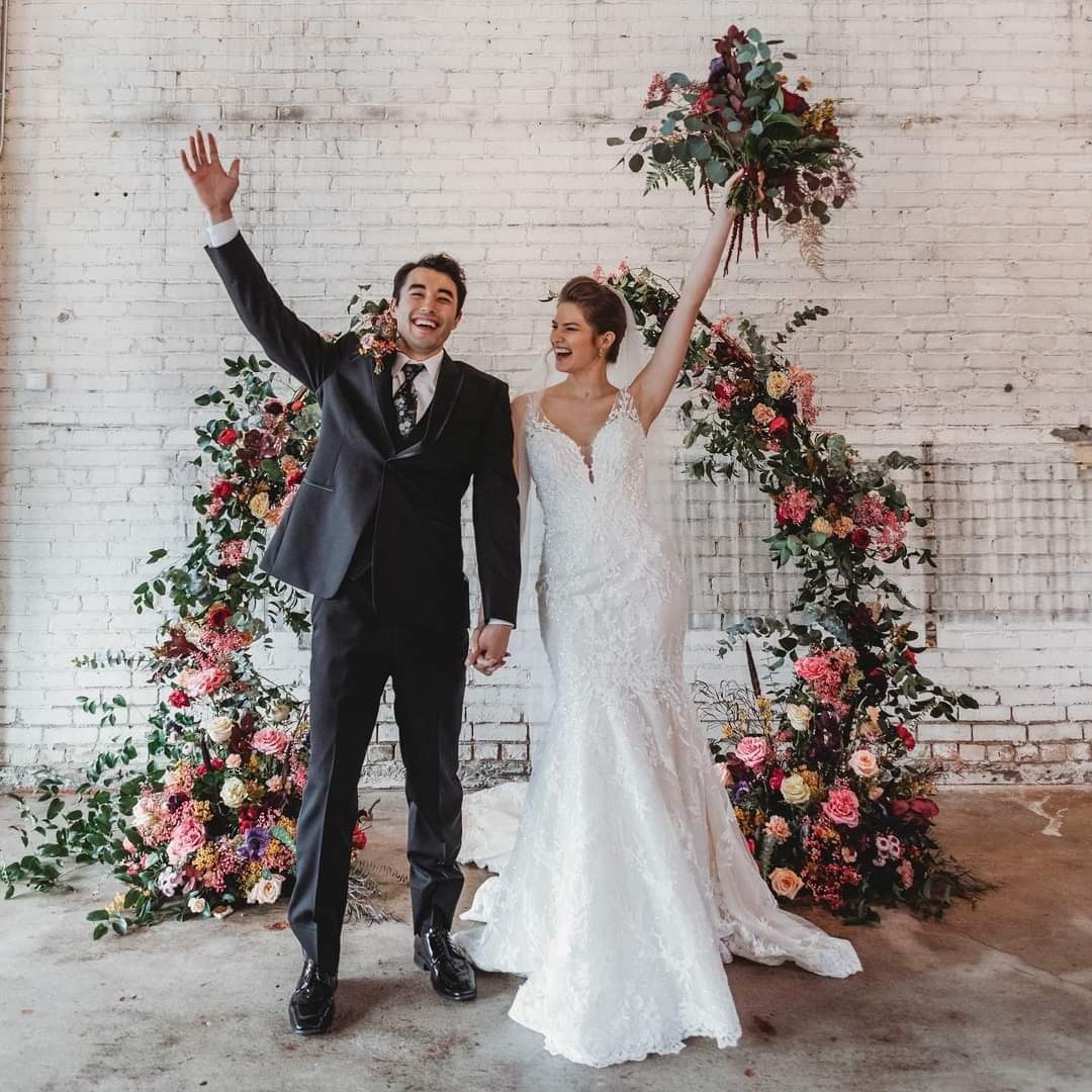 Happy couple in wedding attire holding hands and celebrating with a floral arch and white brick wall background.