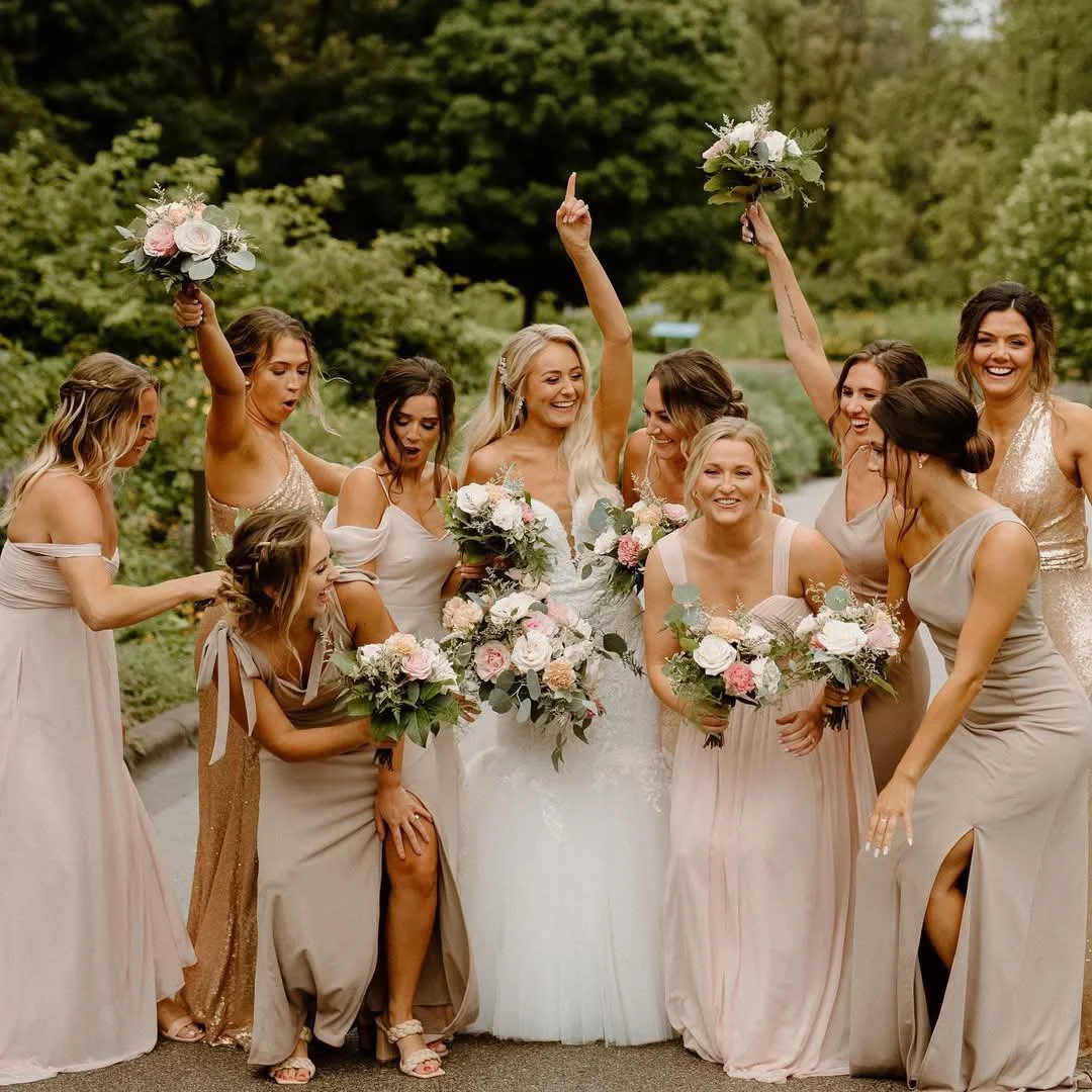 A group of women, including a bride in a white wedding dress and her bridesmaids in beige and champagne dresses, celebrating outdoors. They are holding bouquets of flowers and appear joyful, with some women raising their bouquets and celebrating.