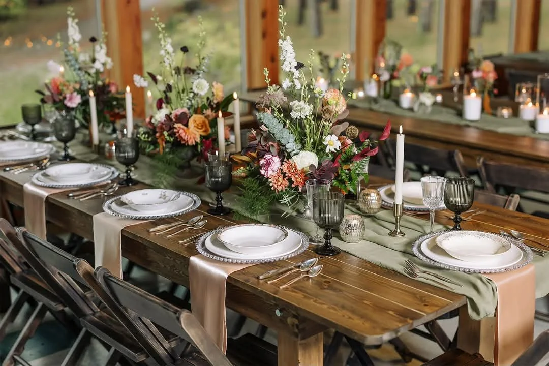 A decorated banquet table with floral centerpieces, candles, glassware, and dinnerware in a rustic setting.