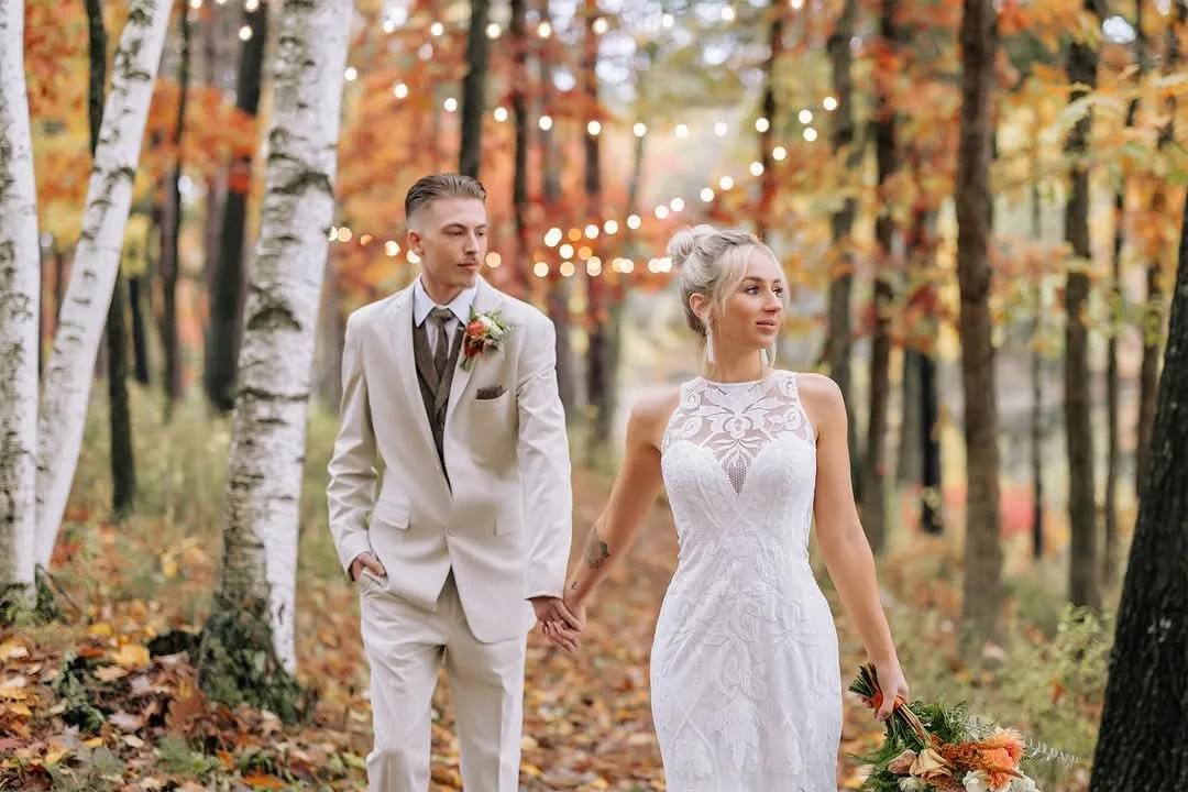 A bride and groom walking hand in hand through an autumn forest decorated with string lights.