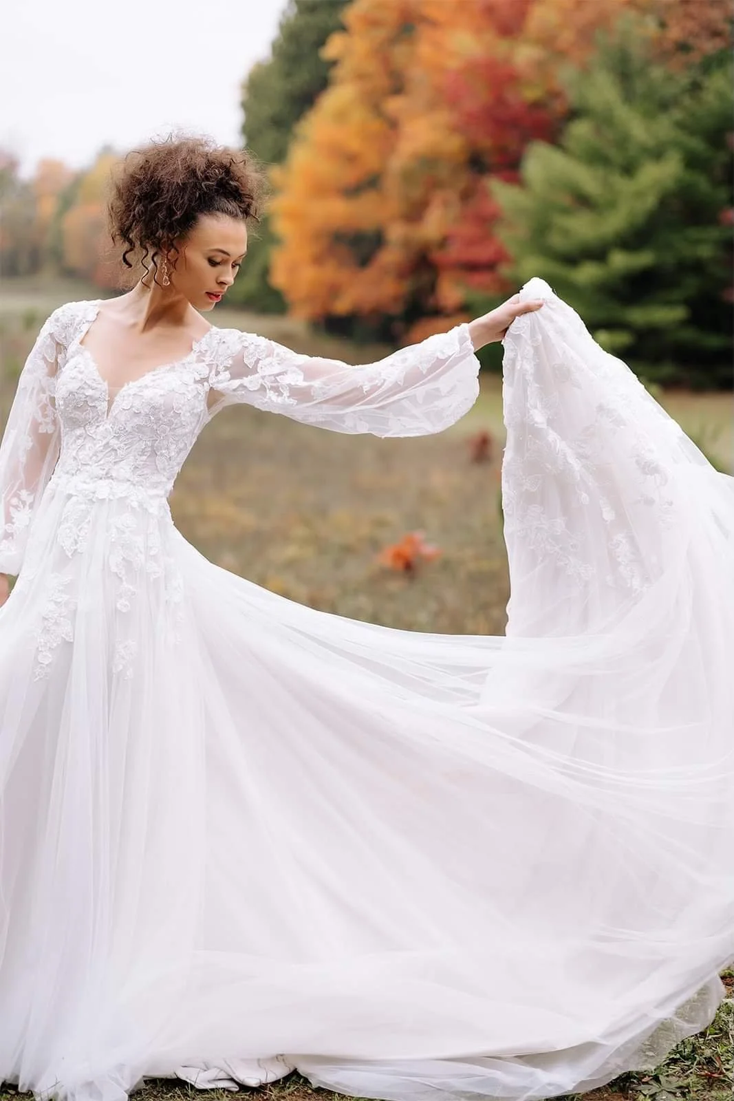 A woman in a white wedding dress with lace details standing outdoors on a fall day with colorful autumn trees in the background.