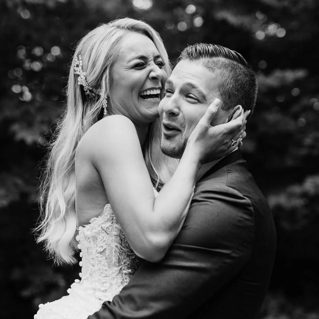 A woman with long blonde hair, wearing a wedding dress, is joyfully hugging a man in a suit. They are outdoors with blurry trees in the background.