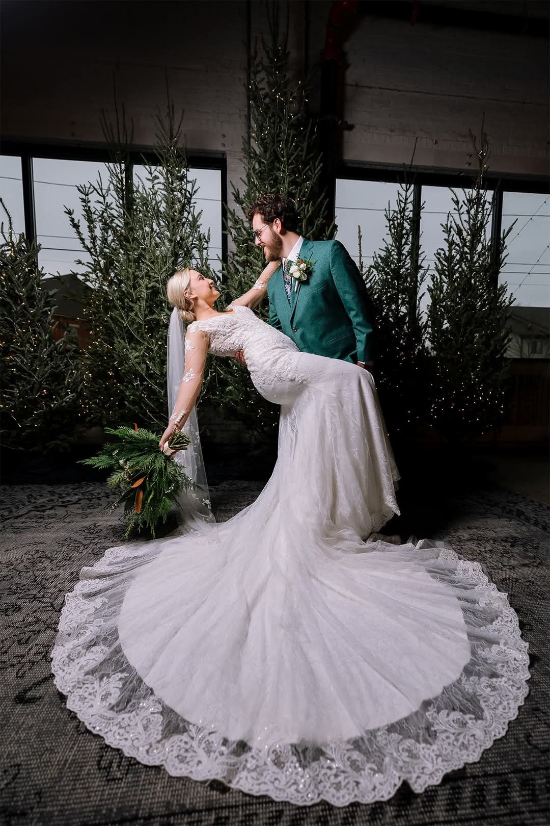 A bride and groom dance together during their wedding in front of decorated Christmas trees, with the bride in a lace wedding dress and the groom in a teal suit, inside a venue with large windows.
