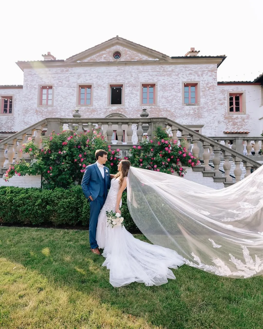 A bride and groom standing on grass in front of a large, white brick mansion with pink window frames and blooming pink roses, dressed in wedding attire, with the bride holding a bouquet and her veil flowing in the wind.
