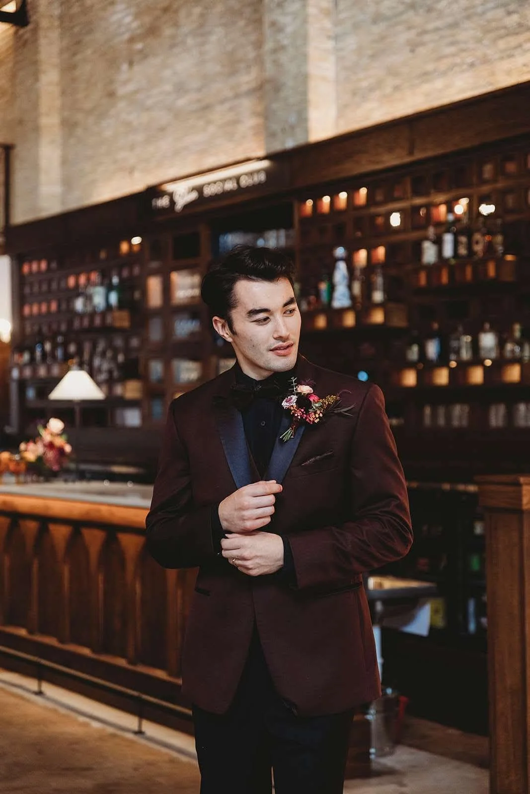 Man in burgundy tuxedo with black bow tie and boutonniere standing in a bar or restaurant, adjusting his boutonniere, with wooden shelves filled with liquor bottles in the background.