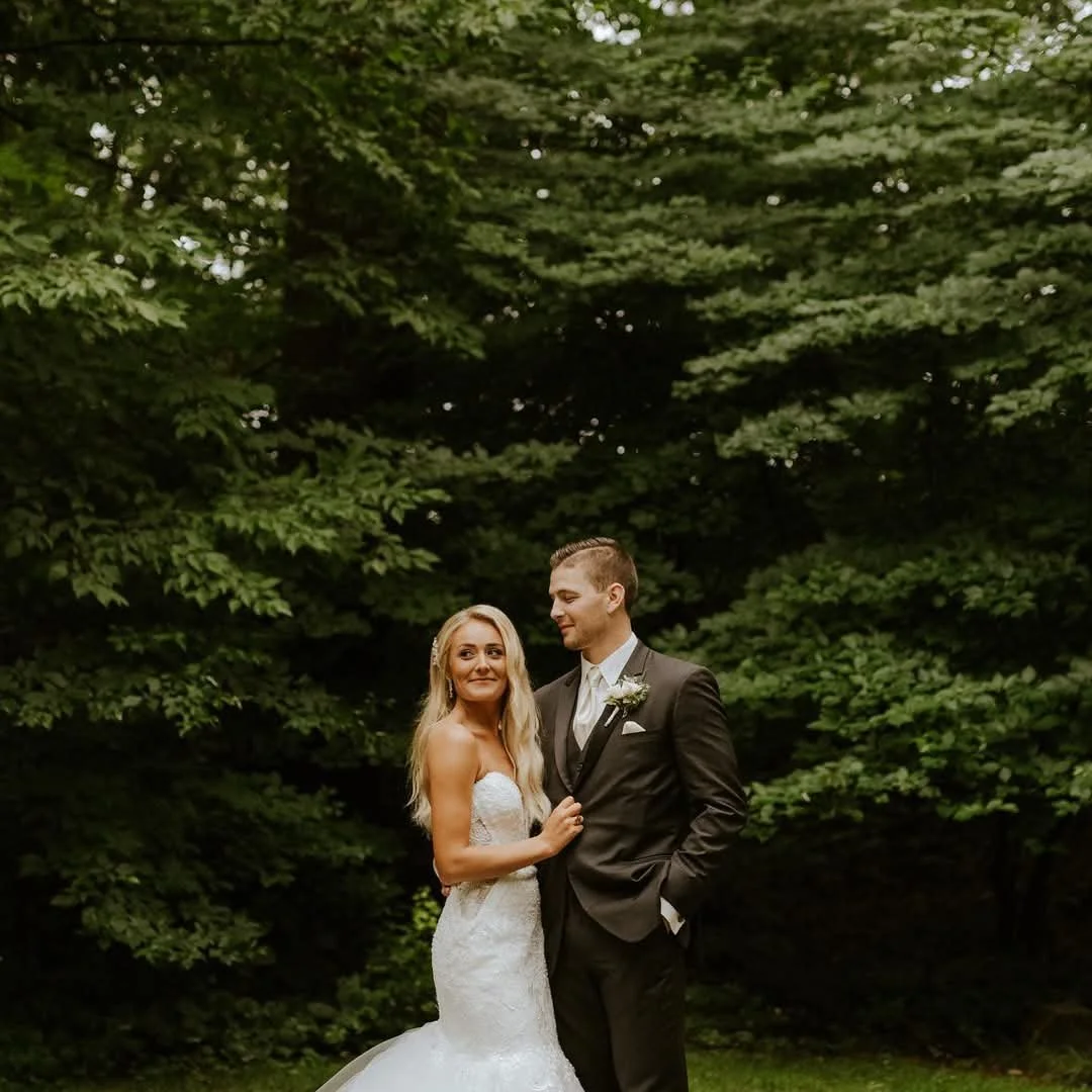 A bride and groom stand together outdoors in front of green trees, dressed in wedding attire. The bride is in a white strapless wedding gown, and the groom is in a dark suit with a white shirt and tie, smiling at each other.