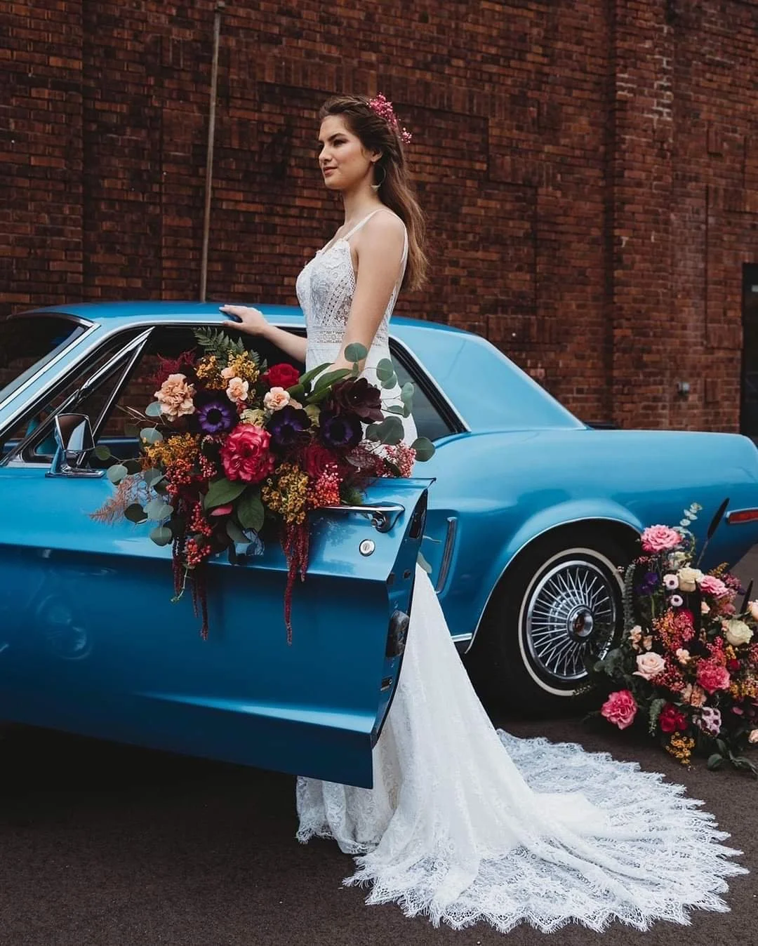A woman in a white wedding dress stands next to a vintage blue car decorated with flowers and holding a large bouquet. Additional flowers are on the ground near the car, with a brick wall in the background.