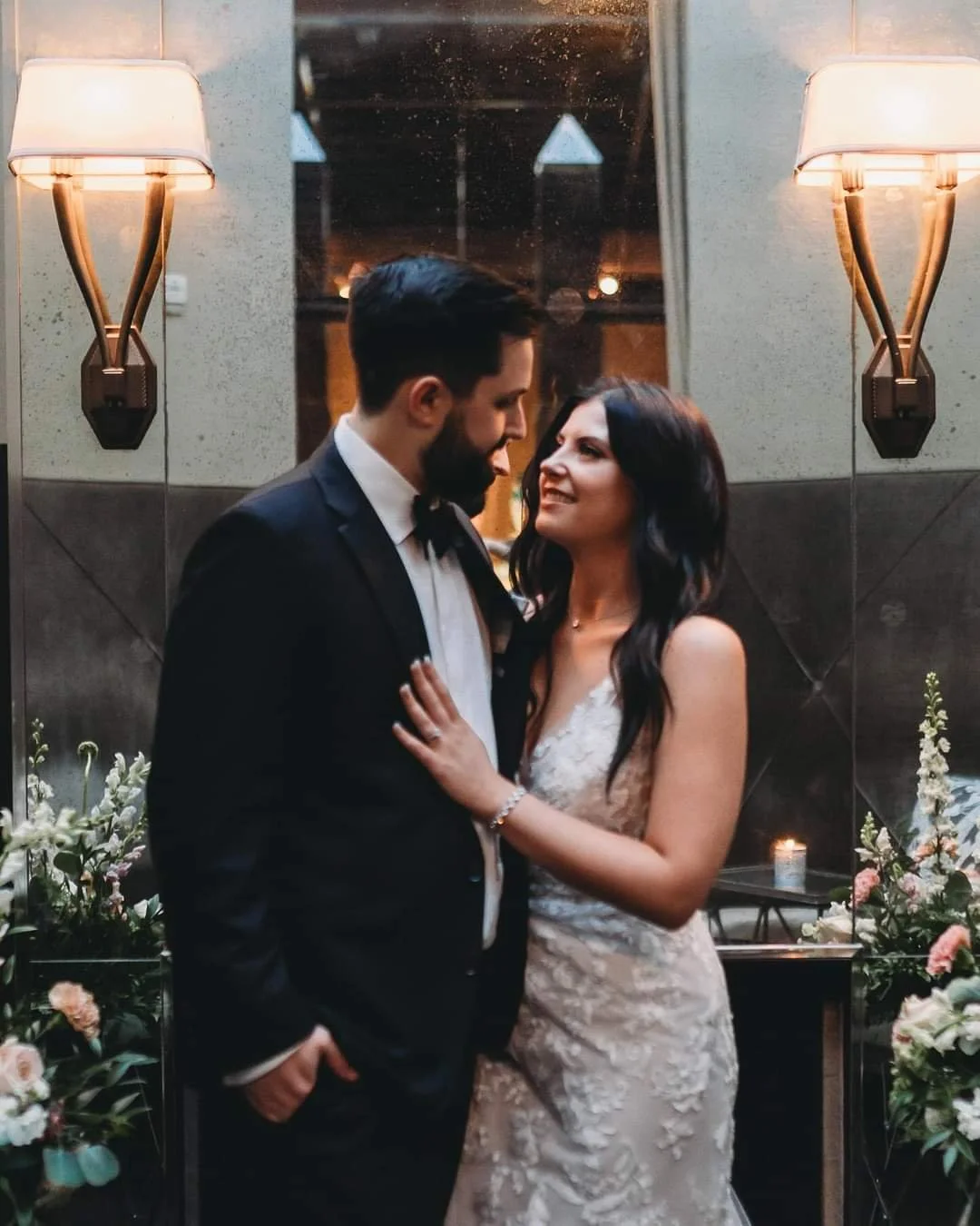 A bride and groom sharing a romantic moment indoors, surrounded by flowers and warm lighting. The groom is wearing a black tuxedo, and the bride is in a white wedding gown.