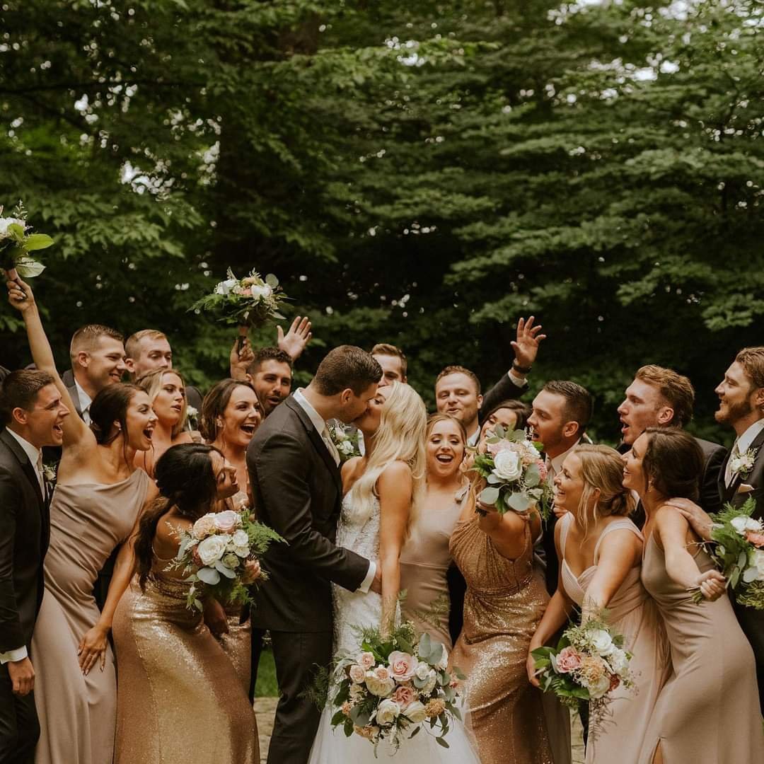 A wedding group photo outdoors with the bride and groom kissing, surrounded by bridesmaids and groomsmen celebrating, holding bouquets, under green trees.