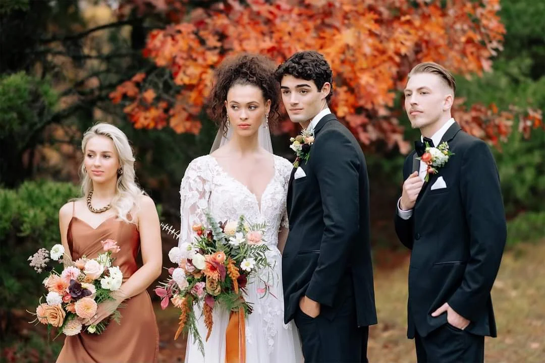 A wedding party of four standing outdoors with autumn orange and green trees in the background. The bride is in a white lace wedding dress holding a floral bouquet, the groom is in a black tuxedo with a boutonniere, a bridesmaid is in a copper-colore