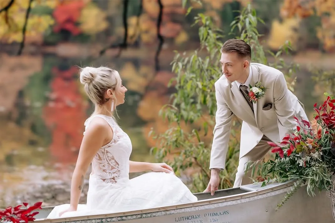 A groom in a white suit helping a bride in a white dress sit in a boat decorated with flowers, by a lake with autumn-colored trees in the background.