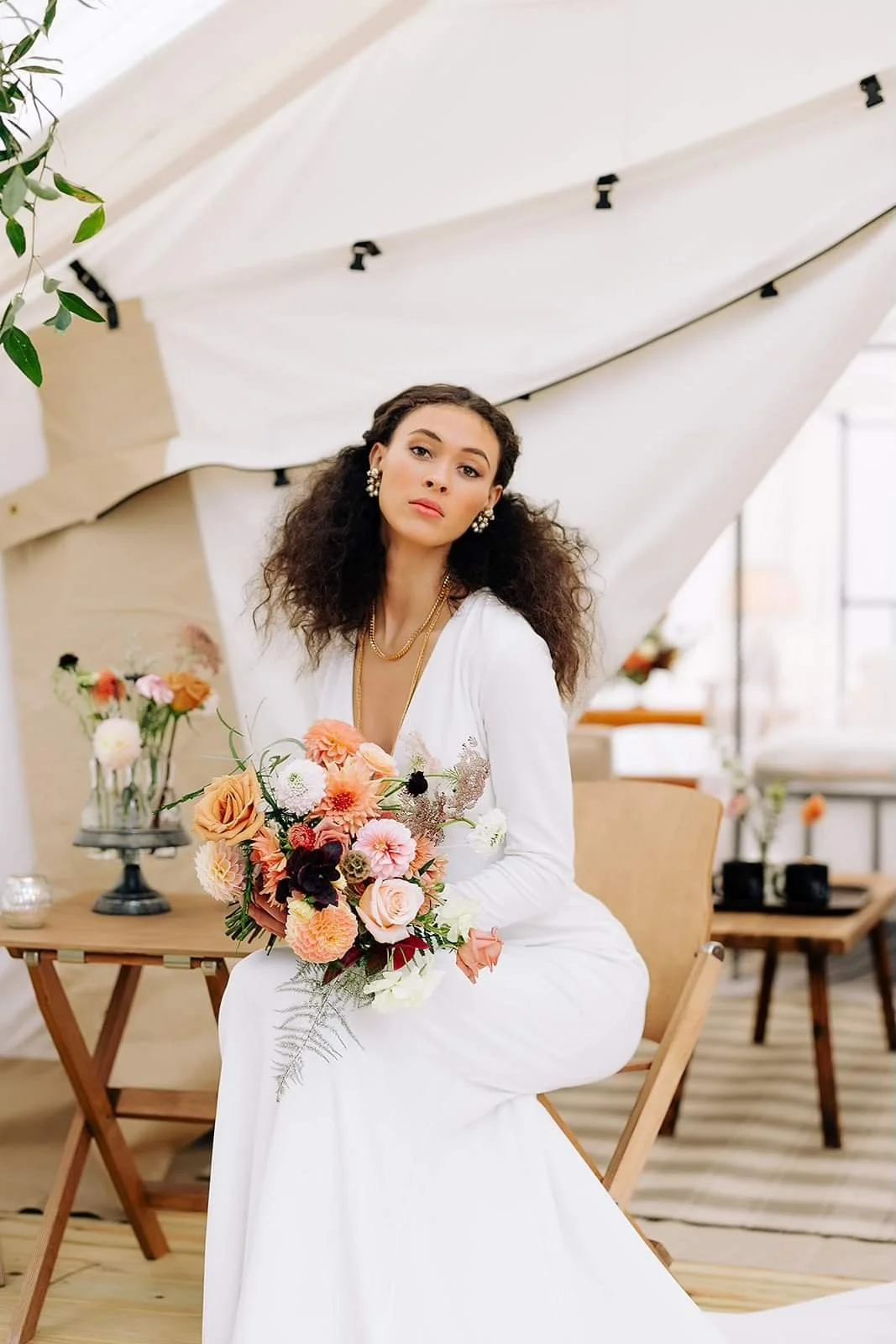 A woman in a white dress sitting on a wooden chair, holding a bouquet of pink, orange, and white flowers, with a neutral expression, in a decorated indoor space with white and wooden elements.