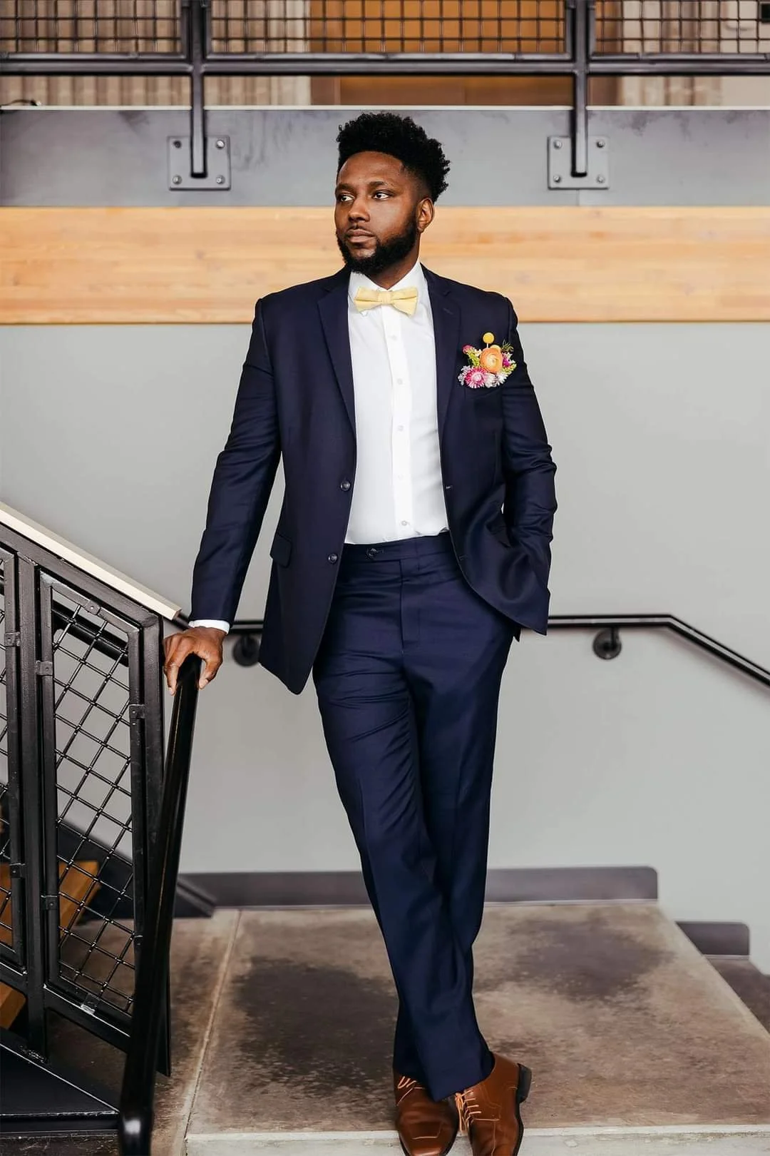 A man in a navy suit, white shirt, and cream bow tie, with a floral boutonniere, standing on a staircase, leaning on the railing, in an indoor setting with wood and metal decor.