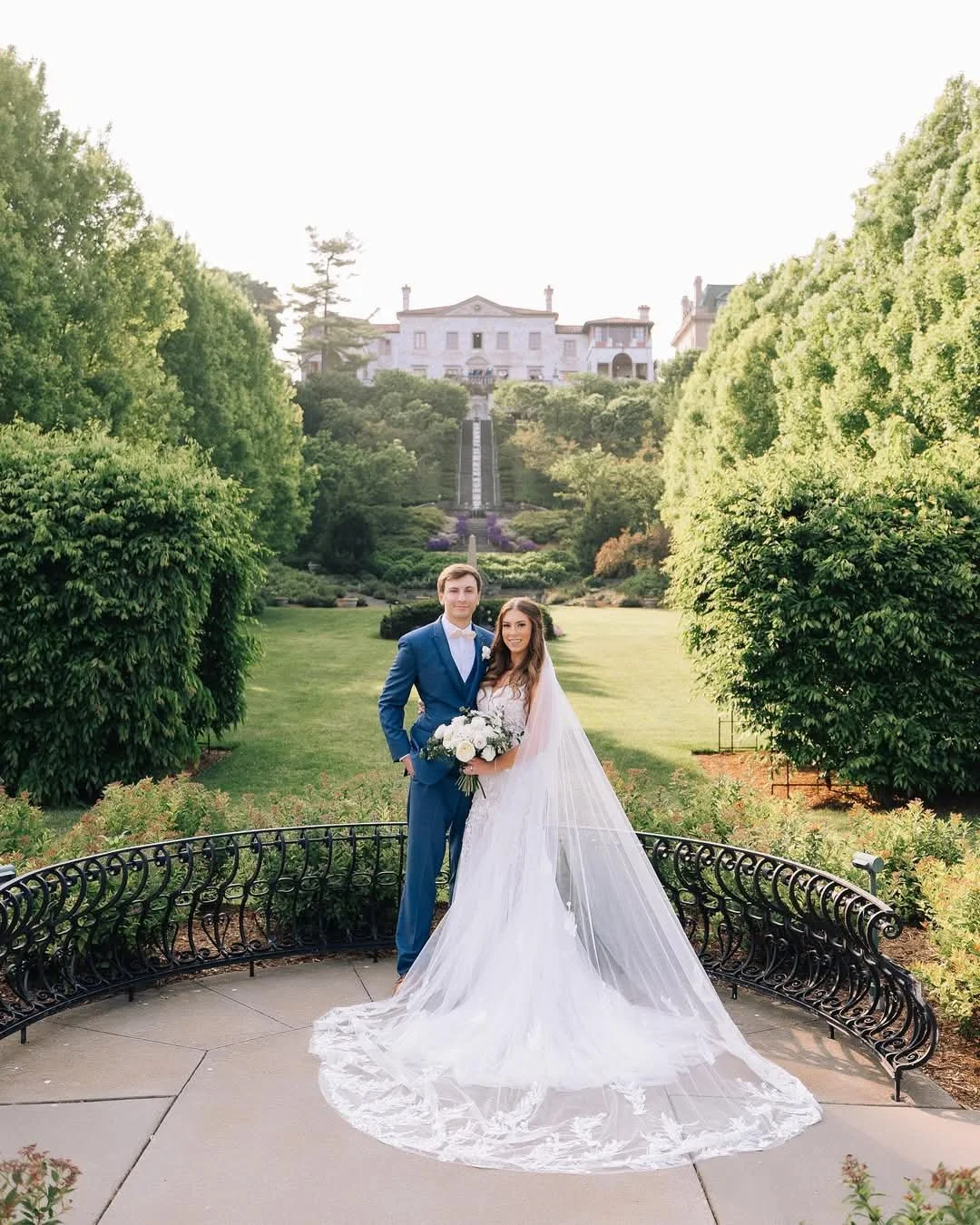 A bride and groom standing on a small curved bench in a garden, with a large mansion-style house in the background. The bride is wearing a white wedding gown with a long train and veil, holding a bouquet of white flowers. The groom is dressed in a bl
