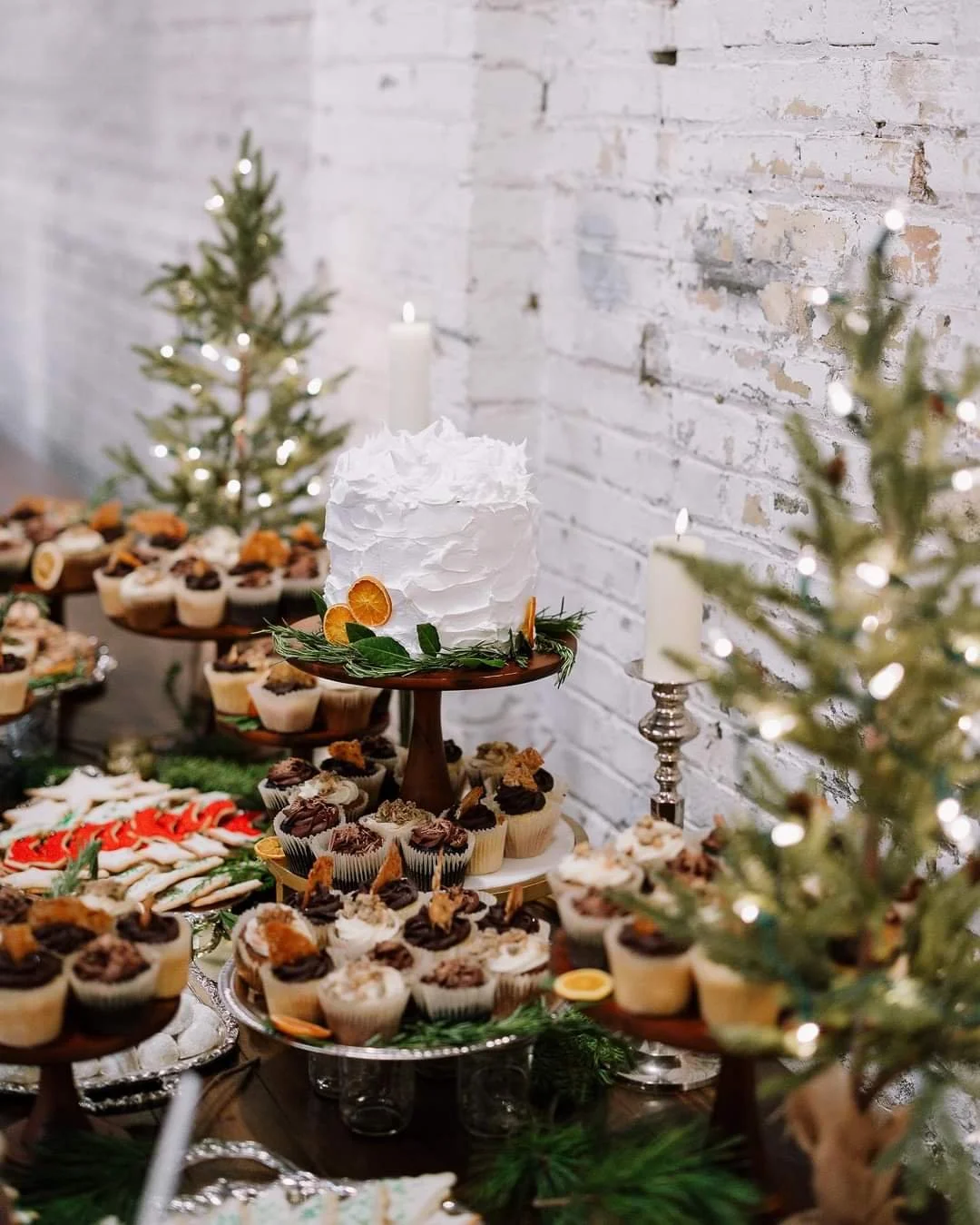 A holiday dessert table with a white frosted cake, cupcakes topped with chocolate and orange slices, Christmas trees, and white candles, set against a white brick wall.