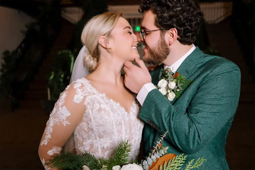 A bride and groom in a close embrace during their wedding, with noses touching and eyes closed, holding a bouquet of greenery and flowers.