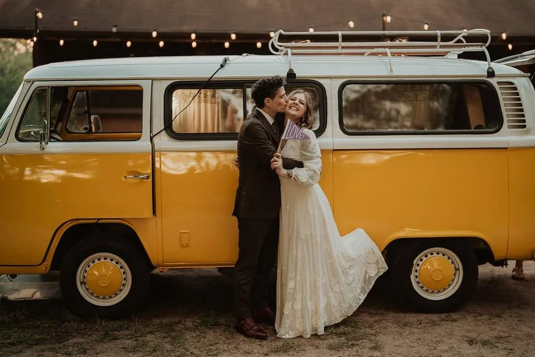 A bride and groom standing in front of a yellow vintage van with a roof rack, at sunset or evening, with string lights overhead. The groom is kissing the bride's cheek, and the bride is smiling while holding a small sign that says "LOVE." The woman w