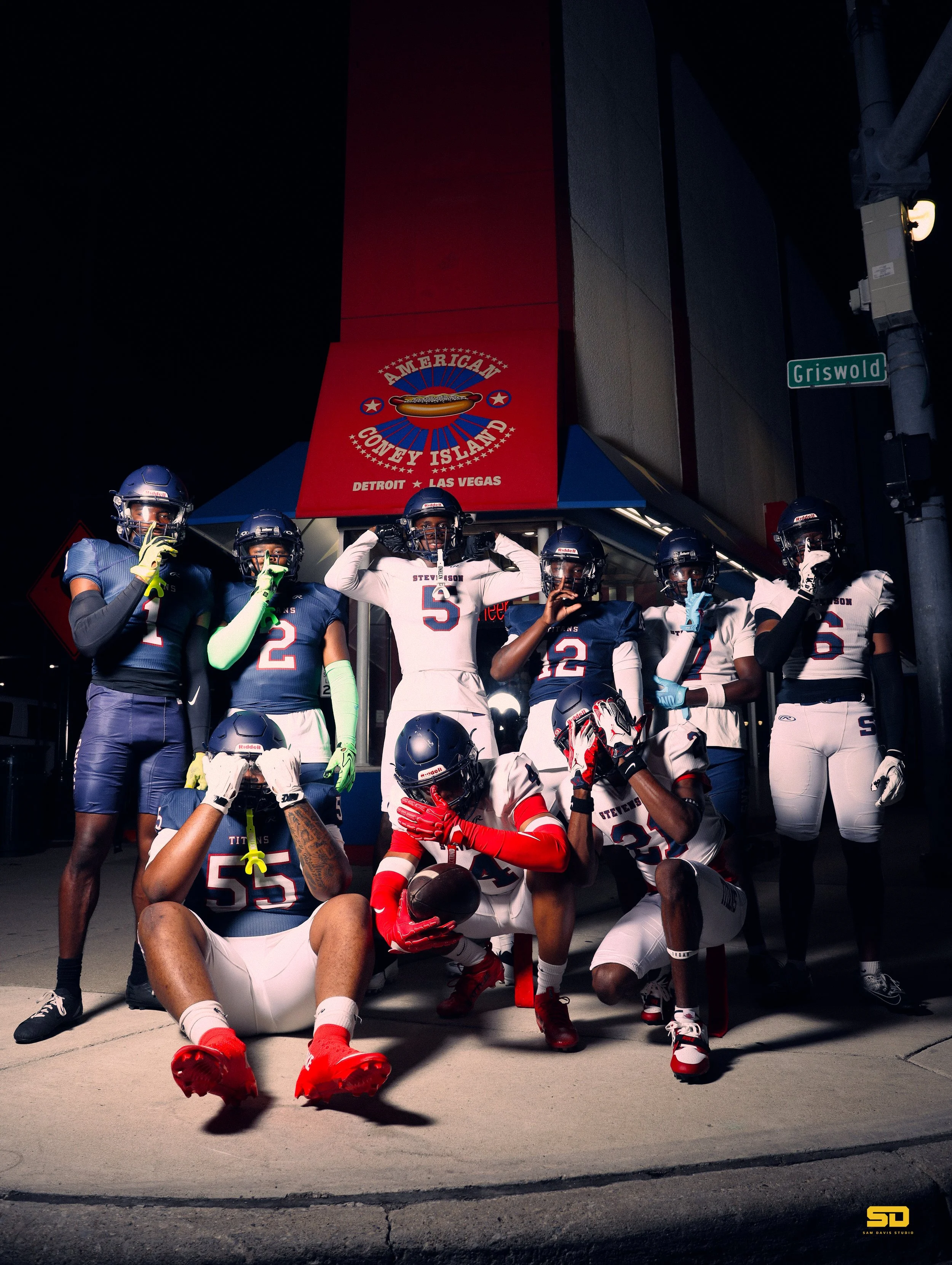 Group of football players in uniforms and helmets posing in front of a building with a red sign that reads 'American County Island' with locations Detroit and Las Vegas. Some players are standing, while others are crouching or kneeling, with one play