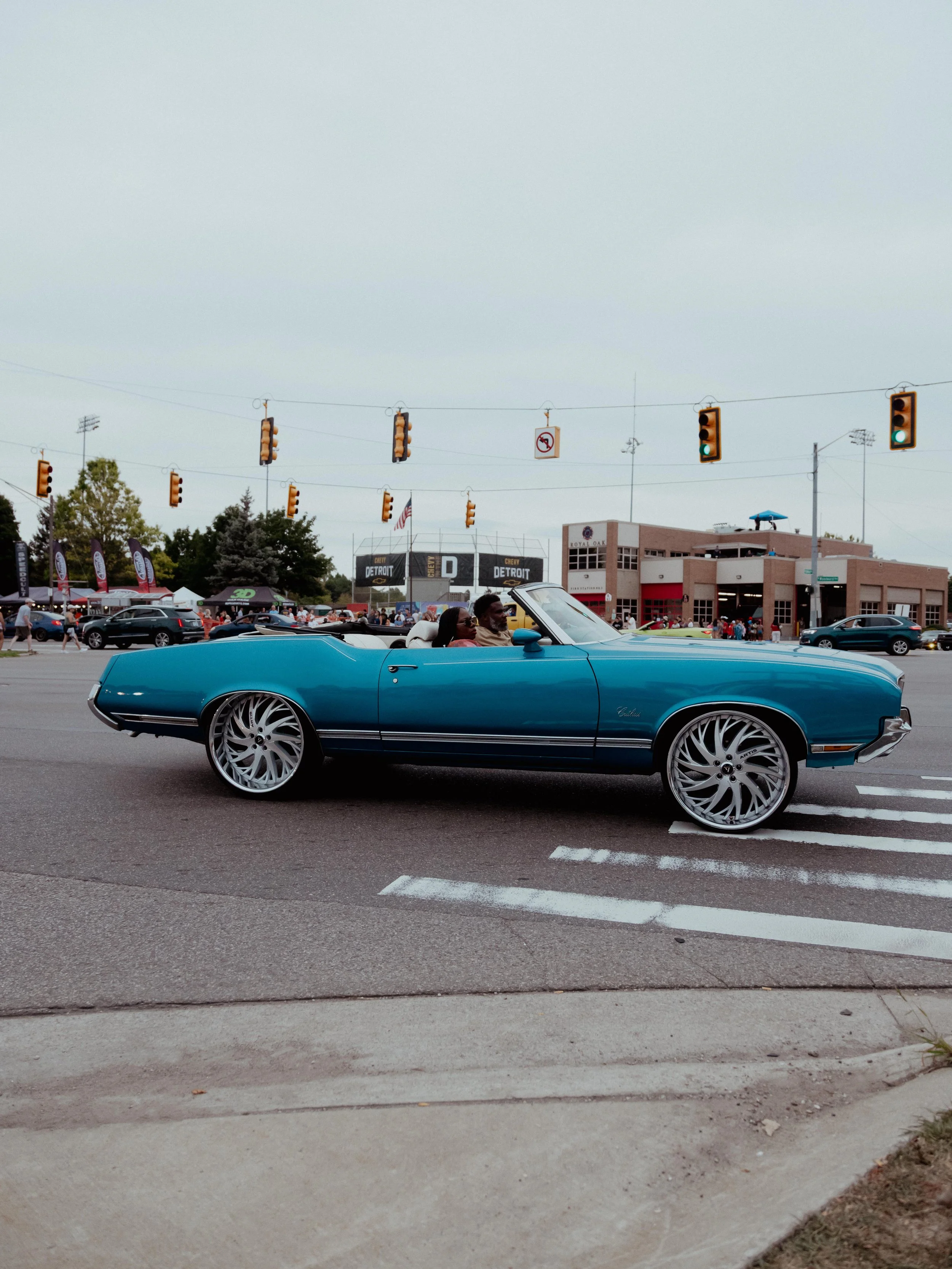 A vintage blue convertible car with large, spiral-style wheels driving down a city street during a car show or parade. The background features a building with a sign that reads 'Royal Oak' and a crowd of people.